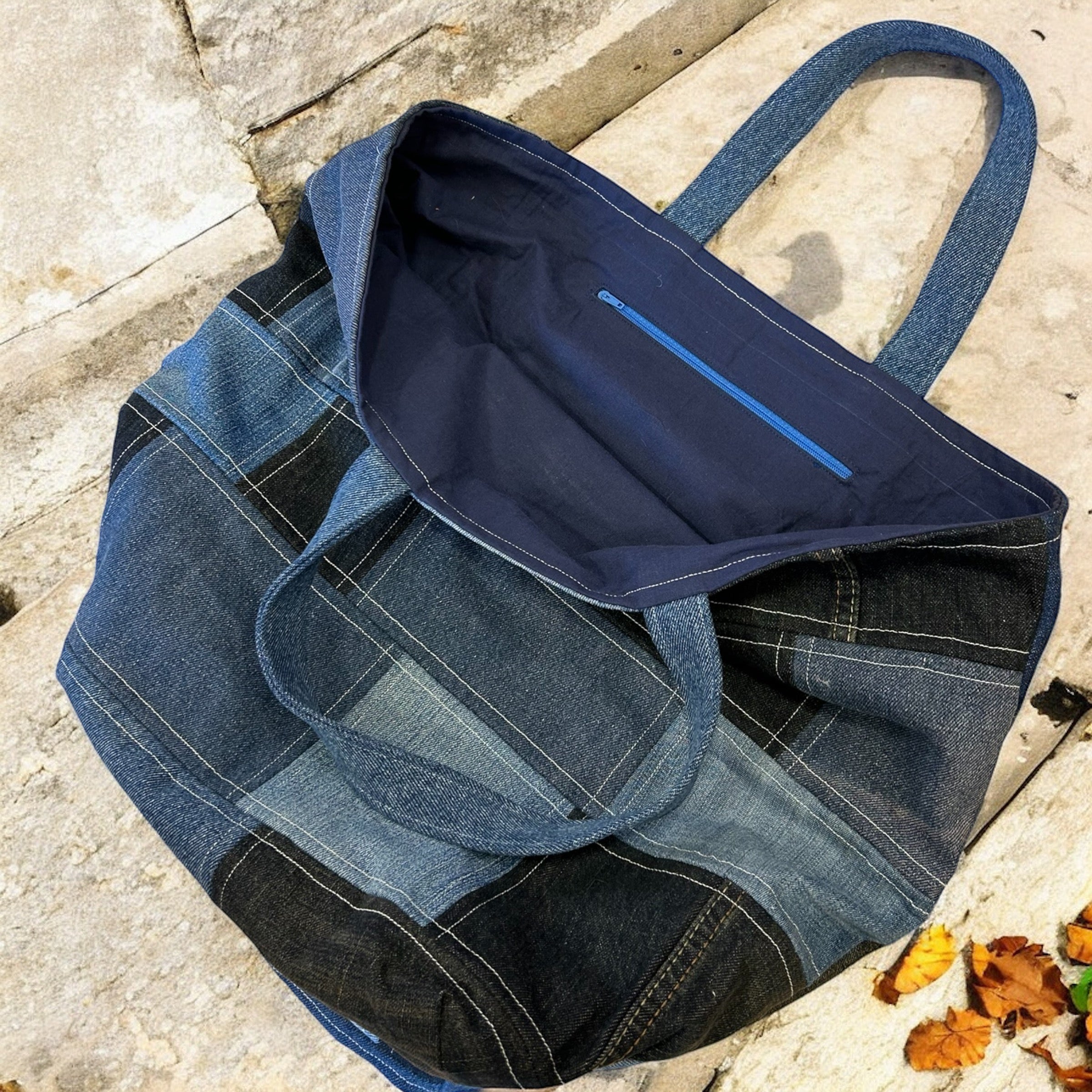 An open view of a large, patchwork denim tote bag, showcasing its dark blue fabric lining with an internal zip pocket. The bag is placed on a stone surface with a few autumn leaves nearby, highlighting the details of its construction and sturdy handles.