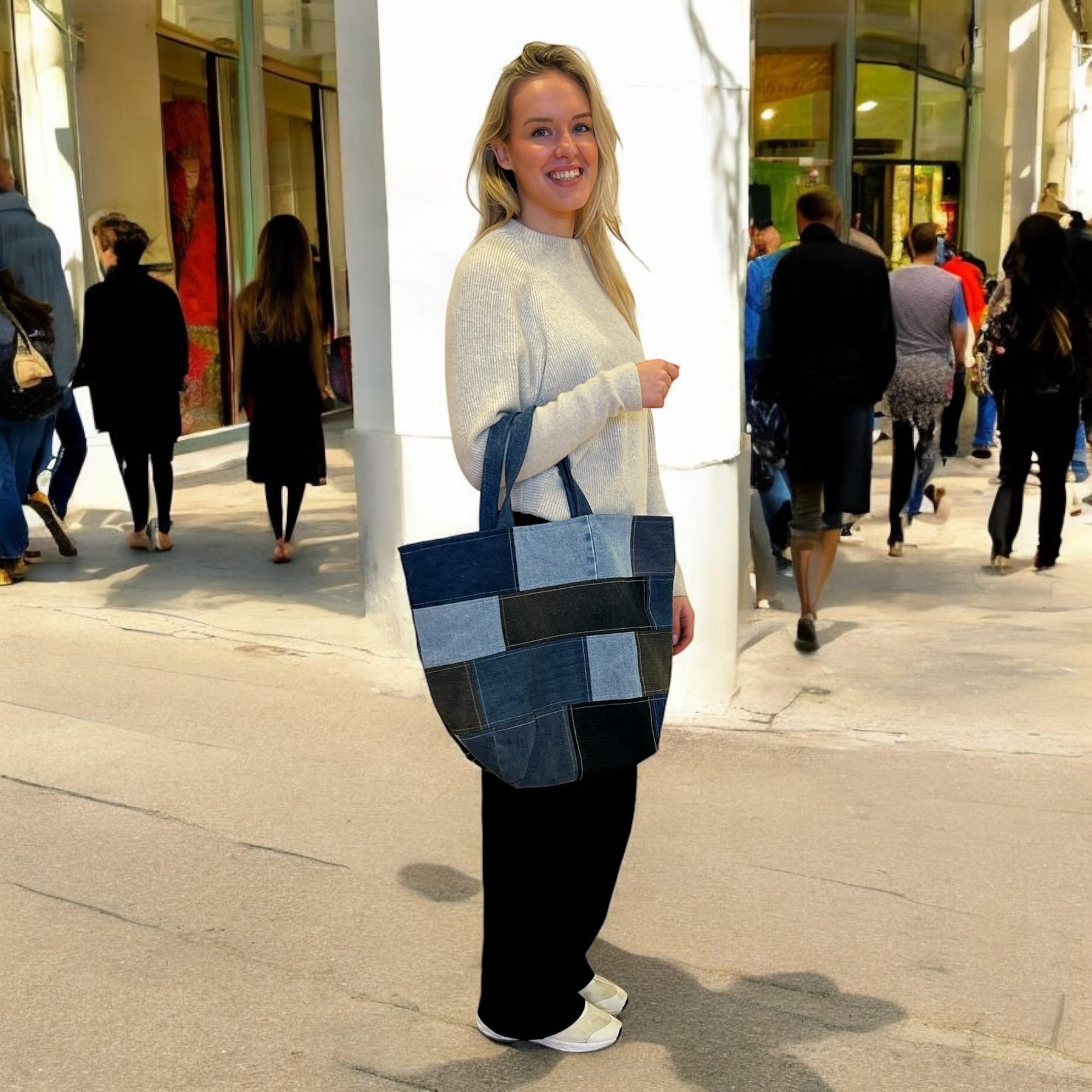 Person standing on a busy street with a large, patchwork tote bag made from various shades of denim. The background shows people walking past shop windows, giving a casual, urban atmosphere. The person is wearing a light sweater, black pants, and white shoes, smiling while holding the denim bag over her arm.