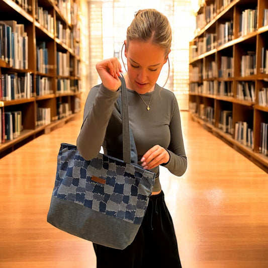 Young woman standing in a brightly lit library, holding a denim patchwork tote bag. The bag features a stylish checkerboard design with contrasting fabric panels and long straps, perfect for carrying books or everyday essentials.