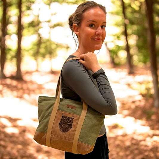 Smiling woman in an outdoor woodland setting carrying a handmade olive green and beige tote bag. The bag features a detailed embroidered owl on the front pocket, long shoulder straps, and a rustic, nature-inspired design that complements the natural surroundings