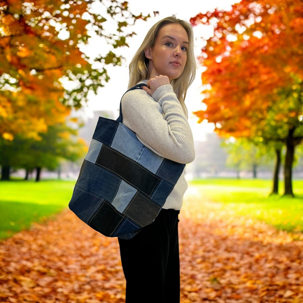 Person standing in a park during autumn, holding a large, patchwork tote bag made from various shades of denim. The background shows trees with vibrant fall foliage in shades of orange, red, and yellow, with fallen leaves scattered on the ground. The person wears a light sweater and black pants, and looks over their shoulder, showcasing the bag.
