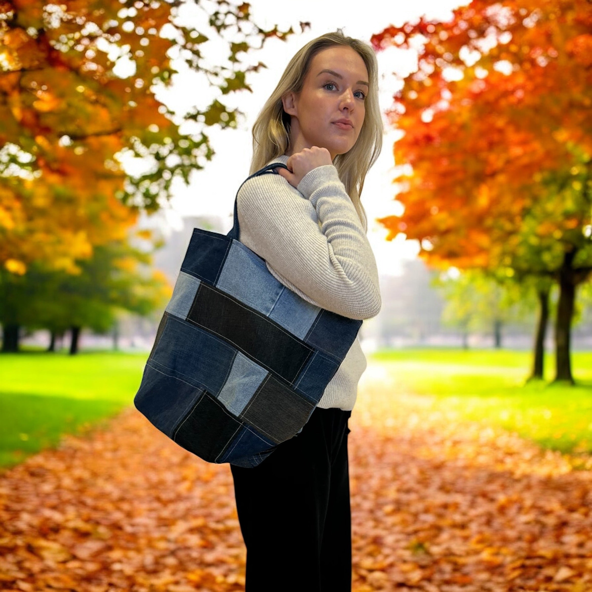 Person standing in a park during autumn, holding a large, patchwork tote bag made from various shades of denim. The background shows trees with vibrant fall foliage in shades of orange, red, and yellow, with fallen leaves scattered on the ground. The person wears a light sweater and black pants, and looks over their shoulder, showcasing the bag.