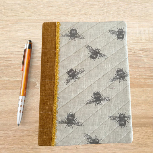 Front view of an A5 notebook cover with black bee print, mustard spine and yellow lace trim, shown with an orange pen on a wooden surface.