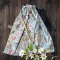 A charming handmade drawstring bag adorned with a delicate pastel floral pattern on a soft cream background. The bag features white rope drawstrings with knotted ends and is displayed on a rustic wooden surface. A sprig of blooming white flowers and green leaves adds a natural, fresh touch to the composition, highlighting the bag's spring-inspired design. A small "Watership Down Crafts" label is visible on the side.