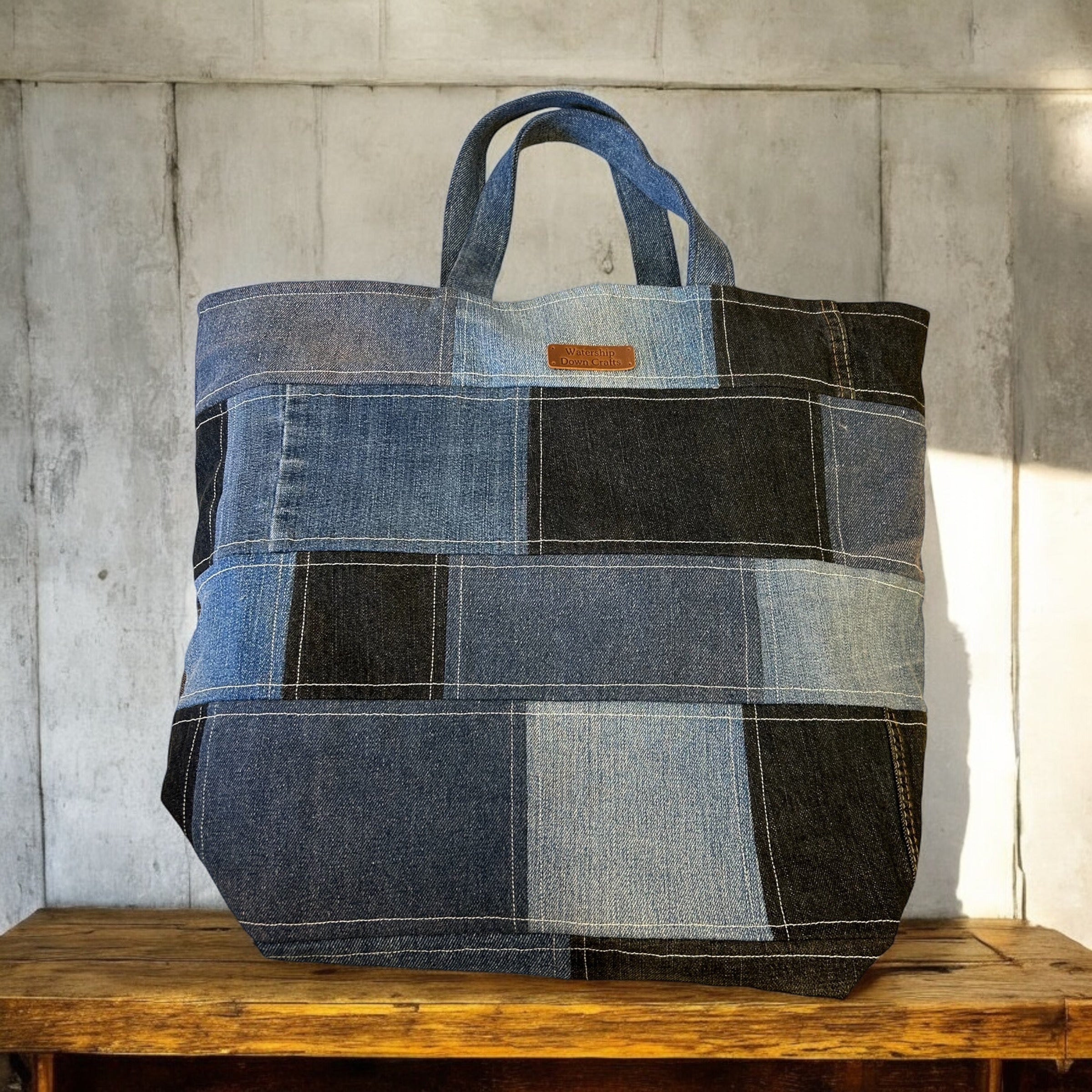 A close-up of a large, denim patchwork tote bag made from various shades of denim fabric, displayed on a rustic wooden surface against a textured, light-colored background. The bag features a woven label that reads 'Watership Down Crafts,' adding a personal touch to the design