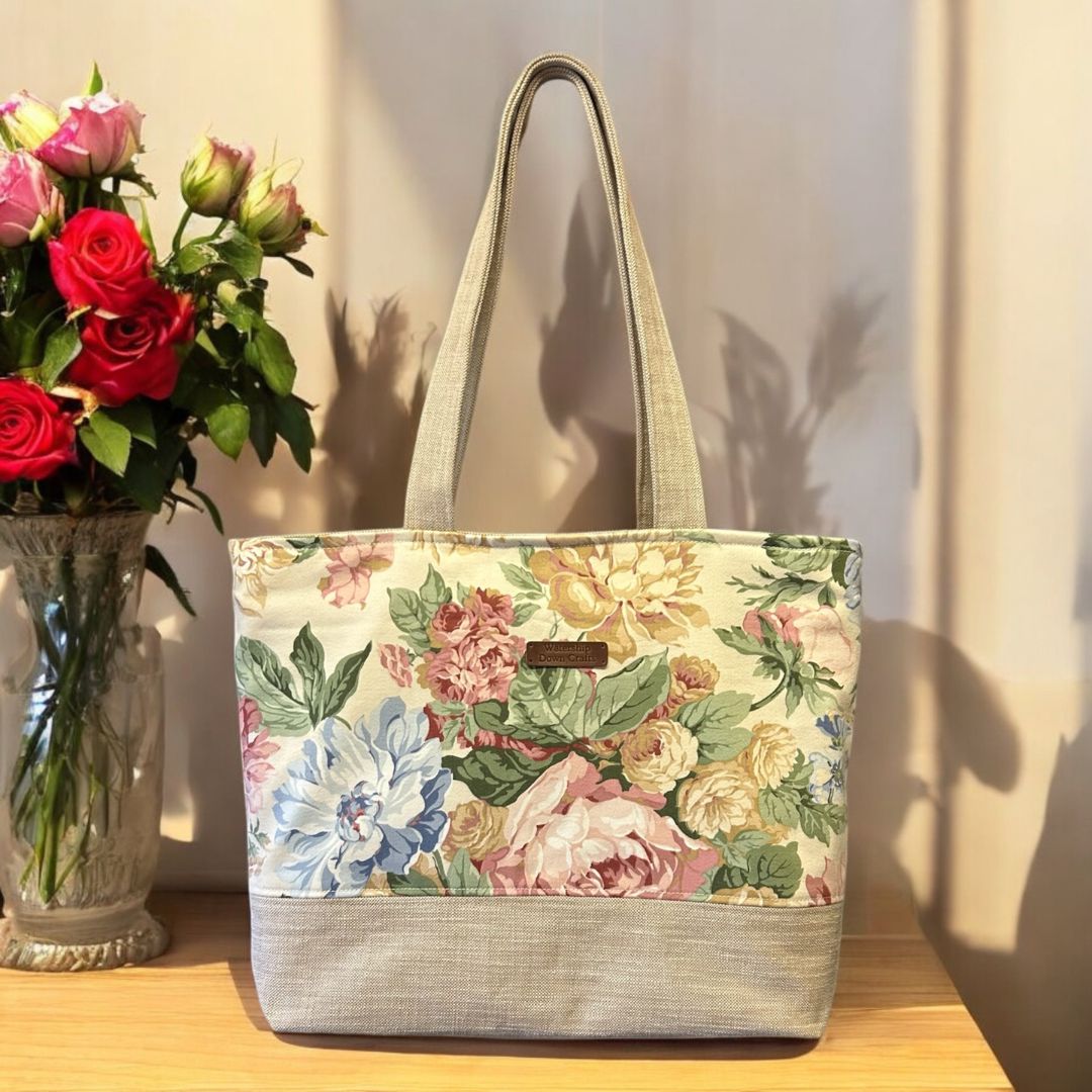 Floral tote bag with pastel pink, yellow, and green flowers on a cream background, displayed on a wooden surface beside a vase of red roses