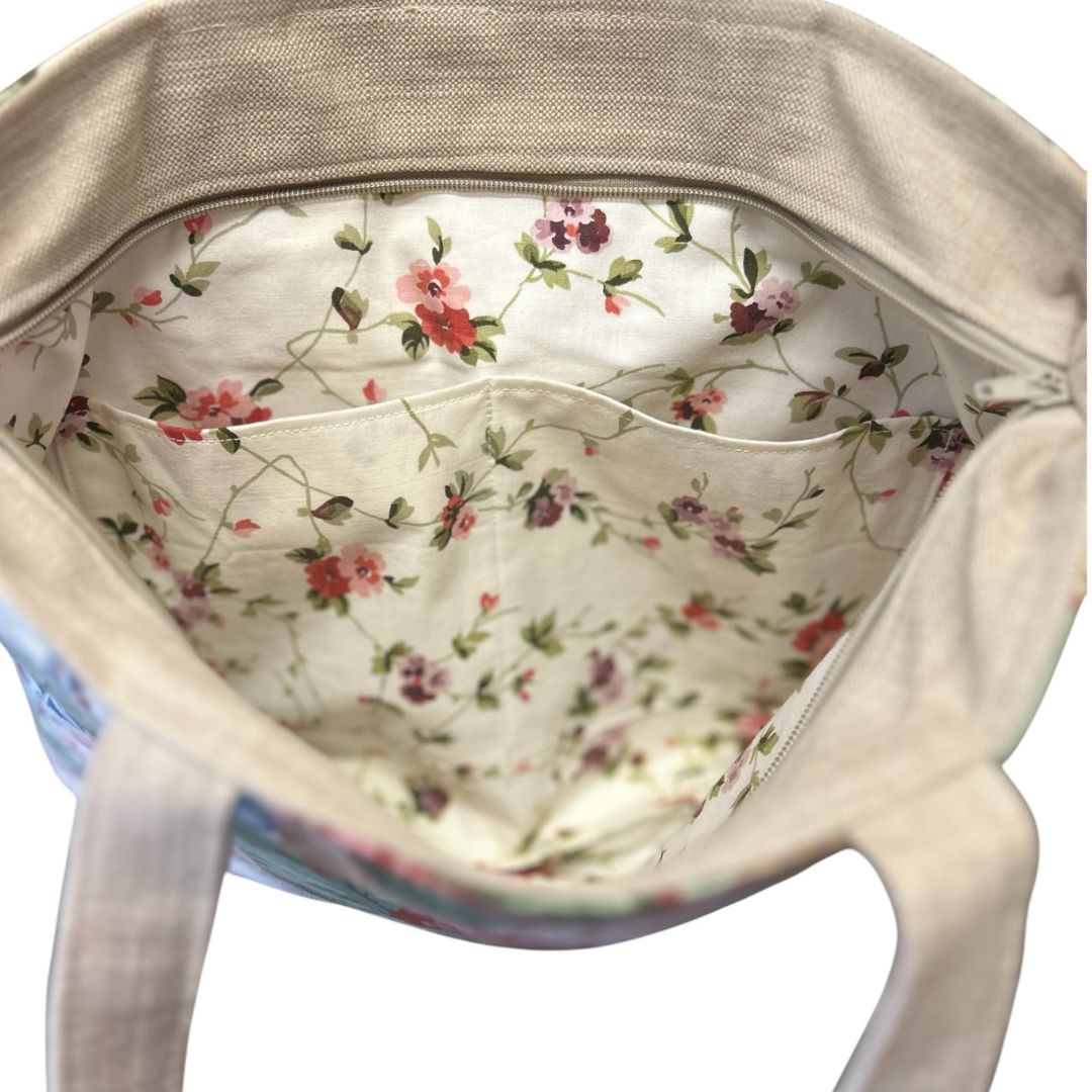Inside view of a floral tote bag showing cream fabric lining with delicate pink and purple flowers, internal slip pocket, and zip closure