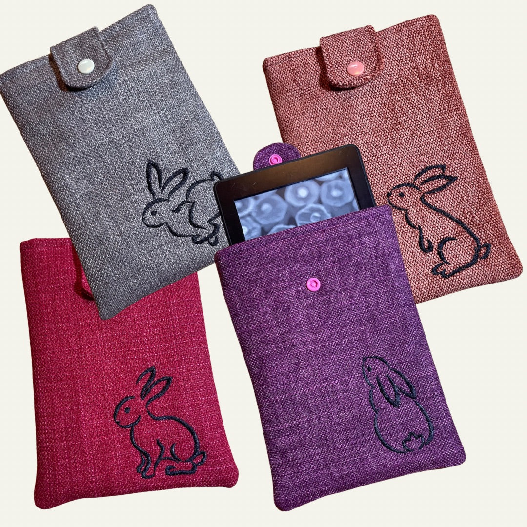 Four handmade fabric Kindle sleeves in gray, purple, orange and pink, each featuring an embroidered rabbit design.