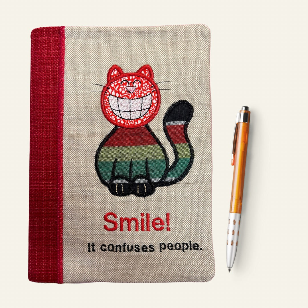 A5 handmade notebook cover with appliqued grinning cat with the embroidered slogan "SMILE it confuses people"