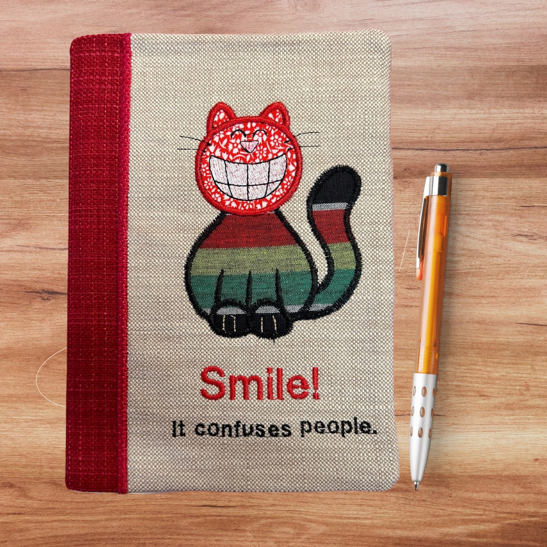 Handmade notebook cover with appliqued grinning cat with embroidered slogan "SMILE it confuses people"