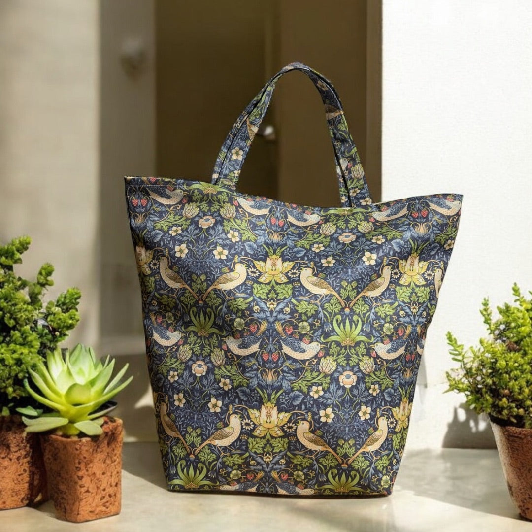 A tote bag with a vibrant William Morris "Strawberry Thief" pattern in blue, green, and beige tones, displayed outdoors on a stone surface. The bag is surrounded by small potted succulents, adding a natural touch to the scene. Sunlight highlights the intricate design of the fabric, showcasing the bag’s elegant and sturdy construction