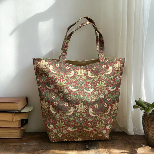 A tote bag with a William Morris "Strawberry Thief" pattern in rich shades of red, green, and beige, displayed on a wooden surface beside a small stack of books and a potted plant. Sunlight streams through a nearby window, casting a soft glow on the bag and highlighting its structured shape and detailed design.