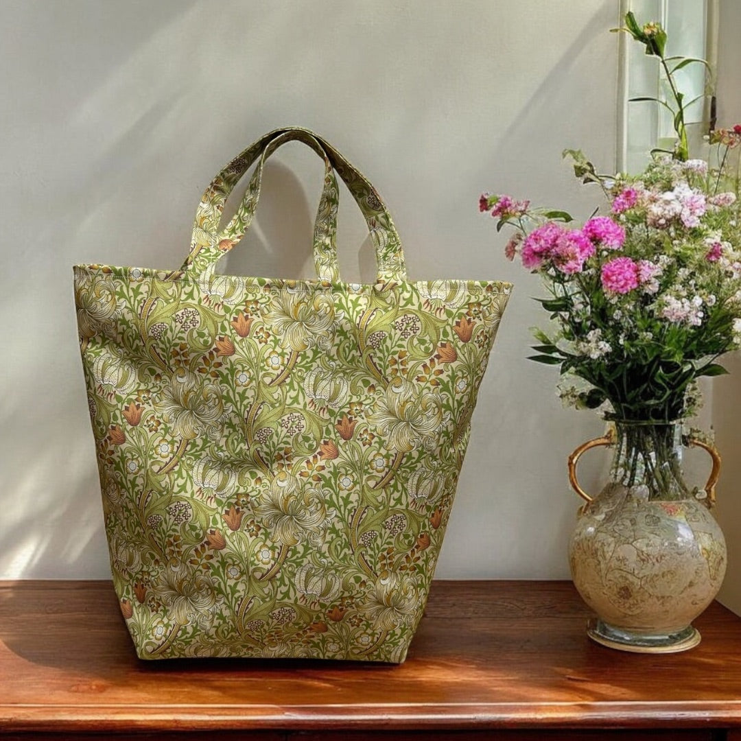 A green William Morris floral-patterned tote bag with intricate botanical designs in shades of white, beige, and orange, displayed on a wooden surface next to a decorative vase filled with pink and white flowers. Soft sunlight illuminates the bag, highlighting its structured shape and sturdy handles, giving a fresh and elegant feel to the scene.