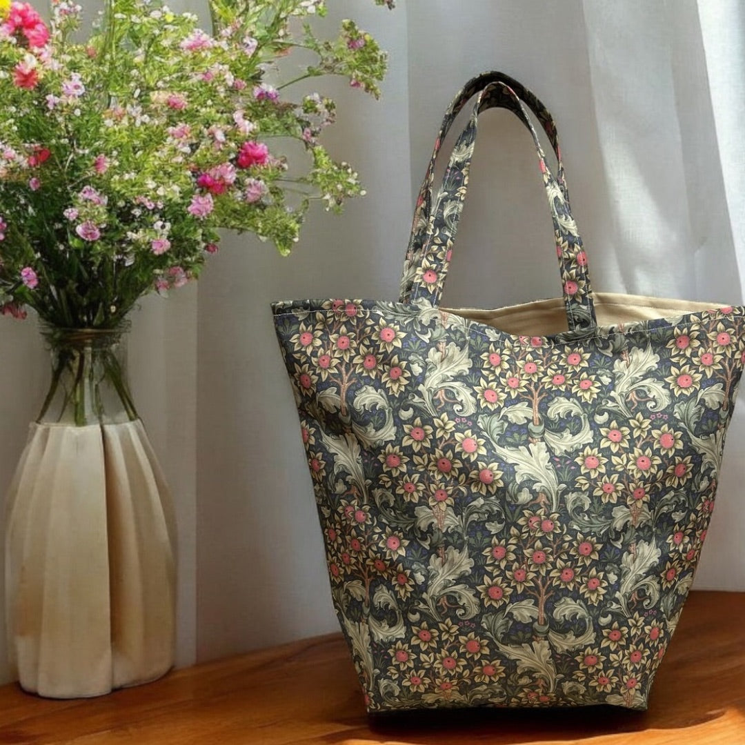 A William Morris floral-patterned tote bag with shades of pink, beige, and green, displayed on a wooden surface beside a vase of fresh pink and white flowers. The bag has a structured shape and matching handles, with the floral design creating a vintage and elegant look. Sunlight filters through, casting a soft glow on the scene