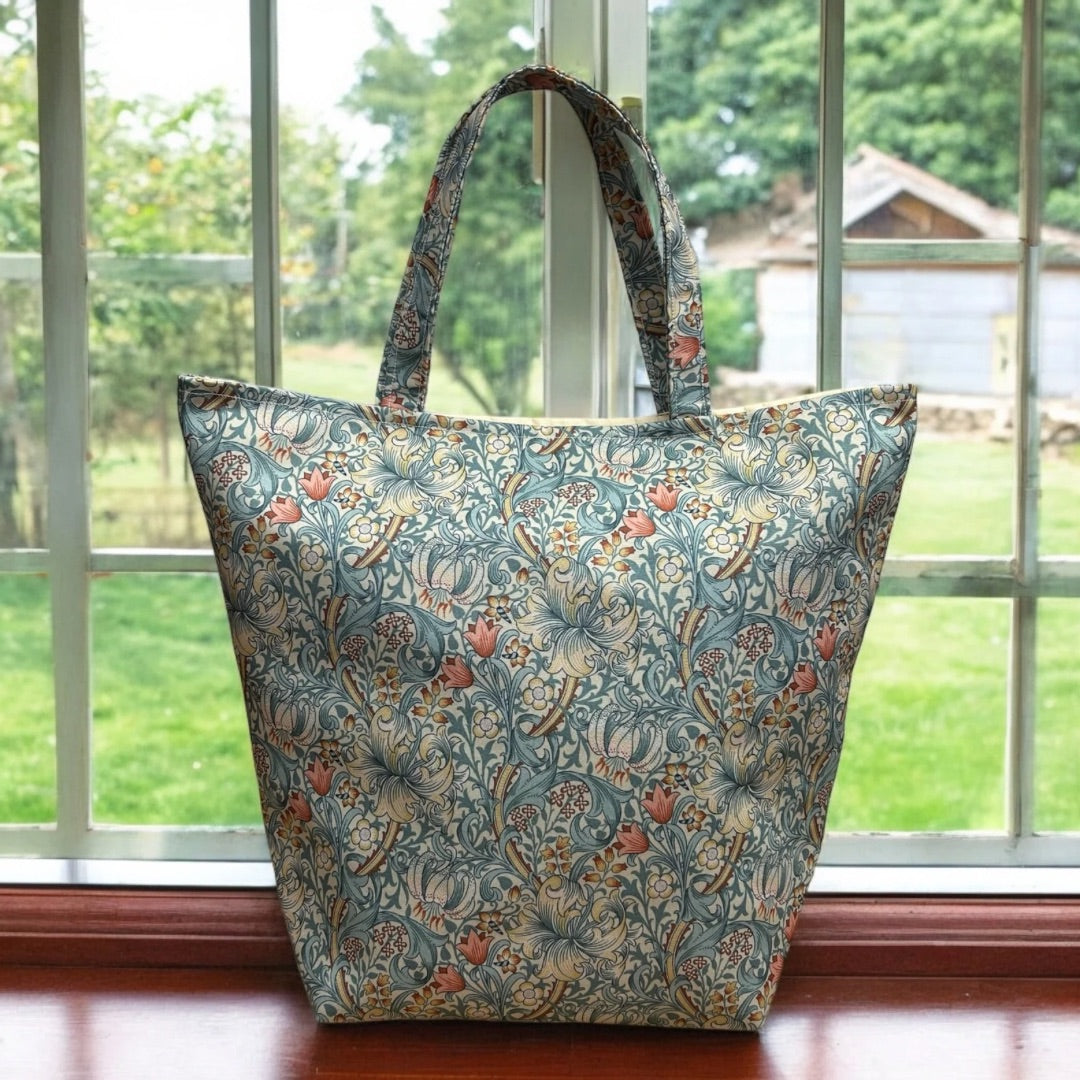 A large tote bag with a floral pattern in soft blue, pink, and beige tones, displayed on a windowsill with a garden view in the background. The bag features sturdy handles and a structured shape, showcasing its detailed, Wiliam Morris designer fabric.