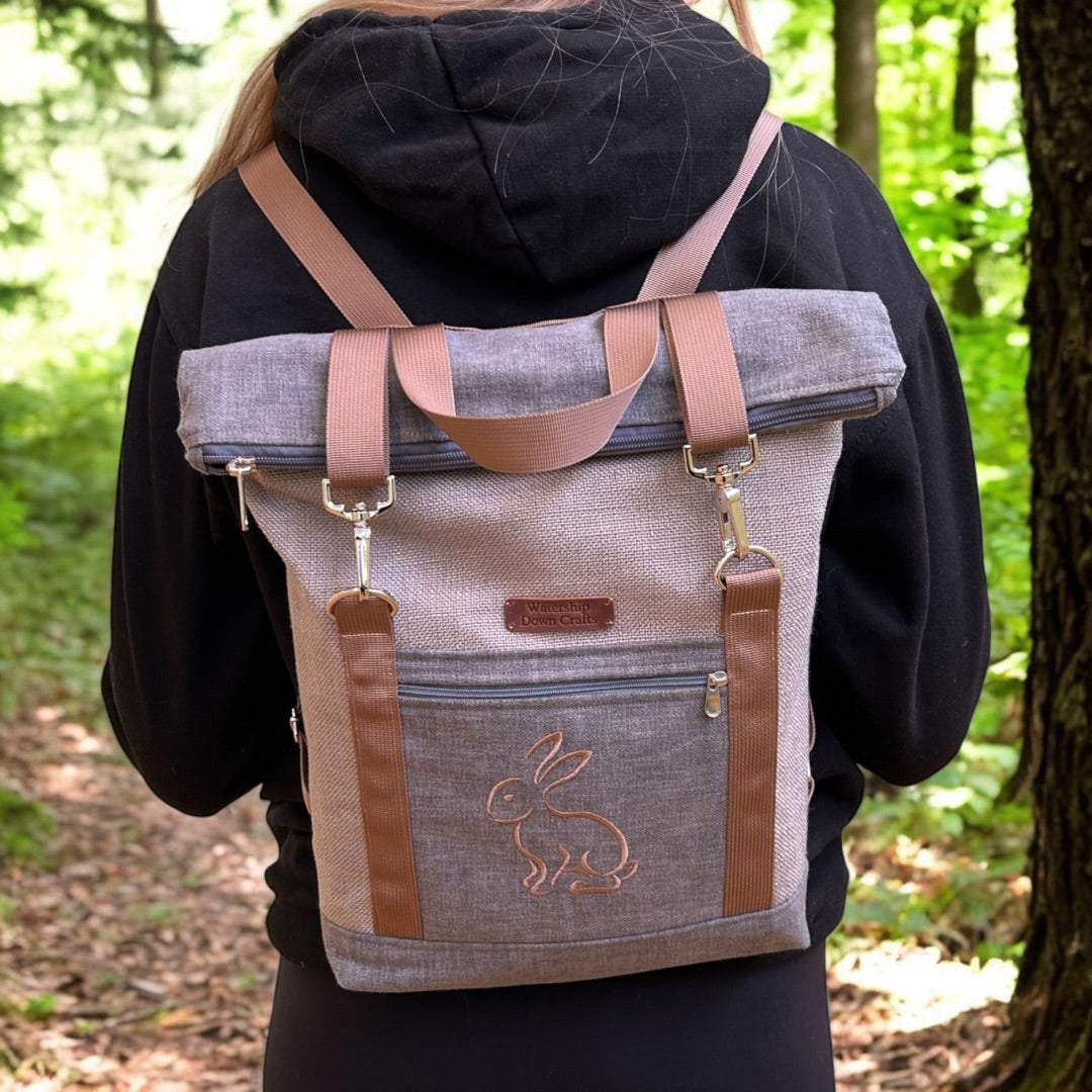 A person wearing a black hoodie is seen from behind, carrying a stylish roll-top backpack in a neutral gray fabric with brown straps. The backpack features a minimalist embroidered outline of a rabbit on the lower front pocket, adding a charming detail. The top of the backpack is rolled down and secured with metal clips attached to the adjustable brown straps. A small leather patch with the "Watership Down Crafts" logo is sewn near the top. The person is standing in a lush forest setting, adding an outdoor,