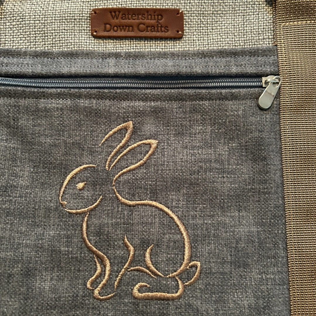 A close-up view of a gray backpack with an embroidered outline of a rabbit on the front pocket. The rabbit design is stitched in a warm tan color, giving it a charming and delicate appearance. Above the zipper of the front pocket, there is a small brown leather patch that reads "Watership Down Crafts." The detailed stitching and earthy tones contribute to the backpack’s handcrafted, nature-inspired aesthetic.