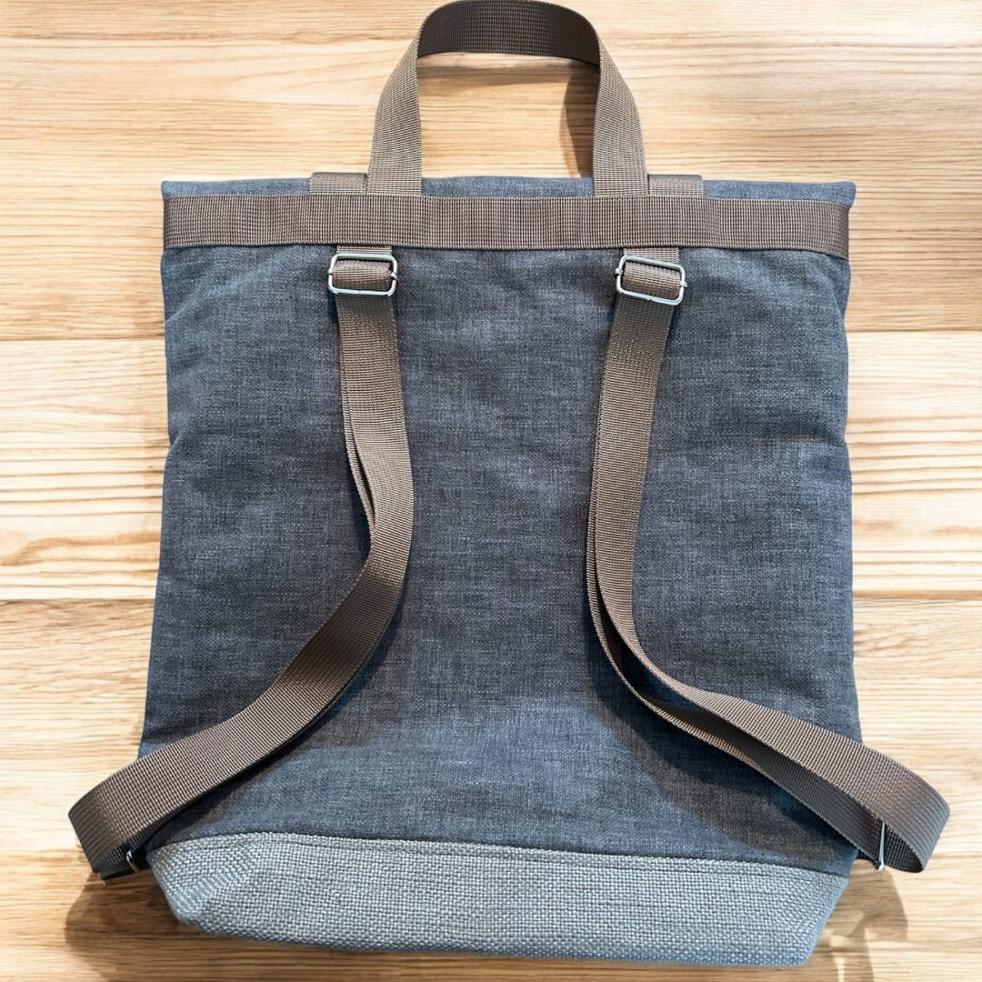 The back view of a gray roll-top backpack laid flat on a wooden surface. The backpack has adjustable brown straps and a matching top handle for carrying. The fabric has a subtle texture, and the bottom of the bag is made of a slightly lighter, durable material. The simplicity of the design and earthy colors give the backpack a modern, minimalist look. The natural wood background complements the bag's rustic aesthetic.