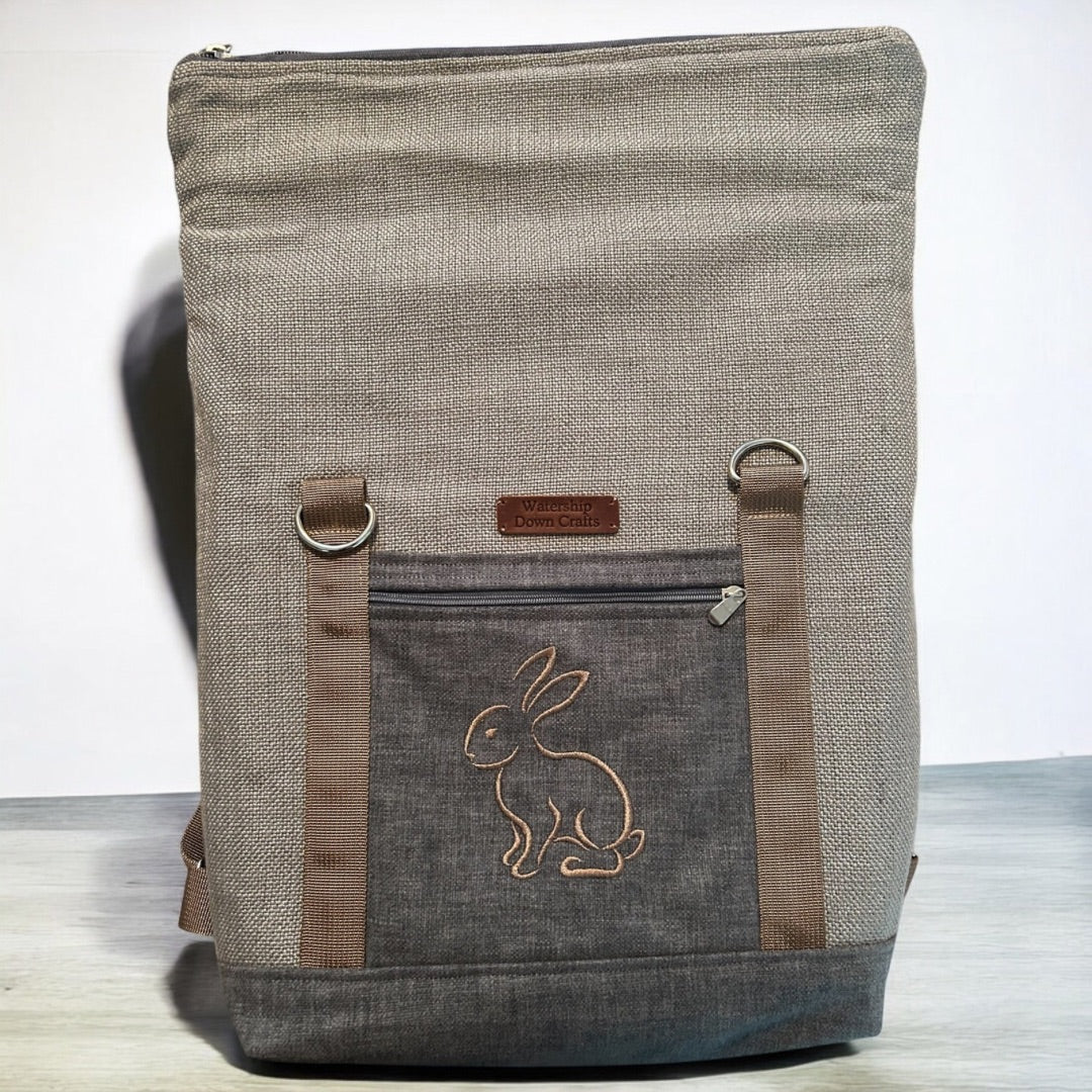 A roll-top backpack made of a textured, neutral gray fabric is displayed upright. The front features a dark gray pocket with an embroidered outline of a rabbit in tan thread, adding a whimsical touch. Above the pocket is a small leather patch with "Watership Down Crafts" embossed on it. The backpack has two brown straps with metal D-rings attached, providing both functionality and style. The minimalist design and earthy tones create a sophisticated yet rustic look, ideal for casual or outdoor use.