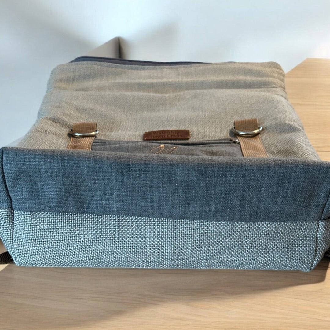 Close-up of the base of a roll-top backpack lying flat, showcasing its durable textured fabric. The base is constructed with a darker, reinforced material to provide added strength and stability. The front of the bag has a smaller pocket with a "Watership Down Crafts" leather patch above it. Two brown straps with metal D-rings are visible, adding functional and aesthetic details. The blend of gray tones and textured fabrics gives the backpack a rugged, handcrafted appearance, ideal for everyday use.