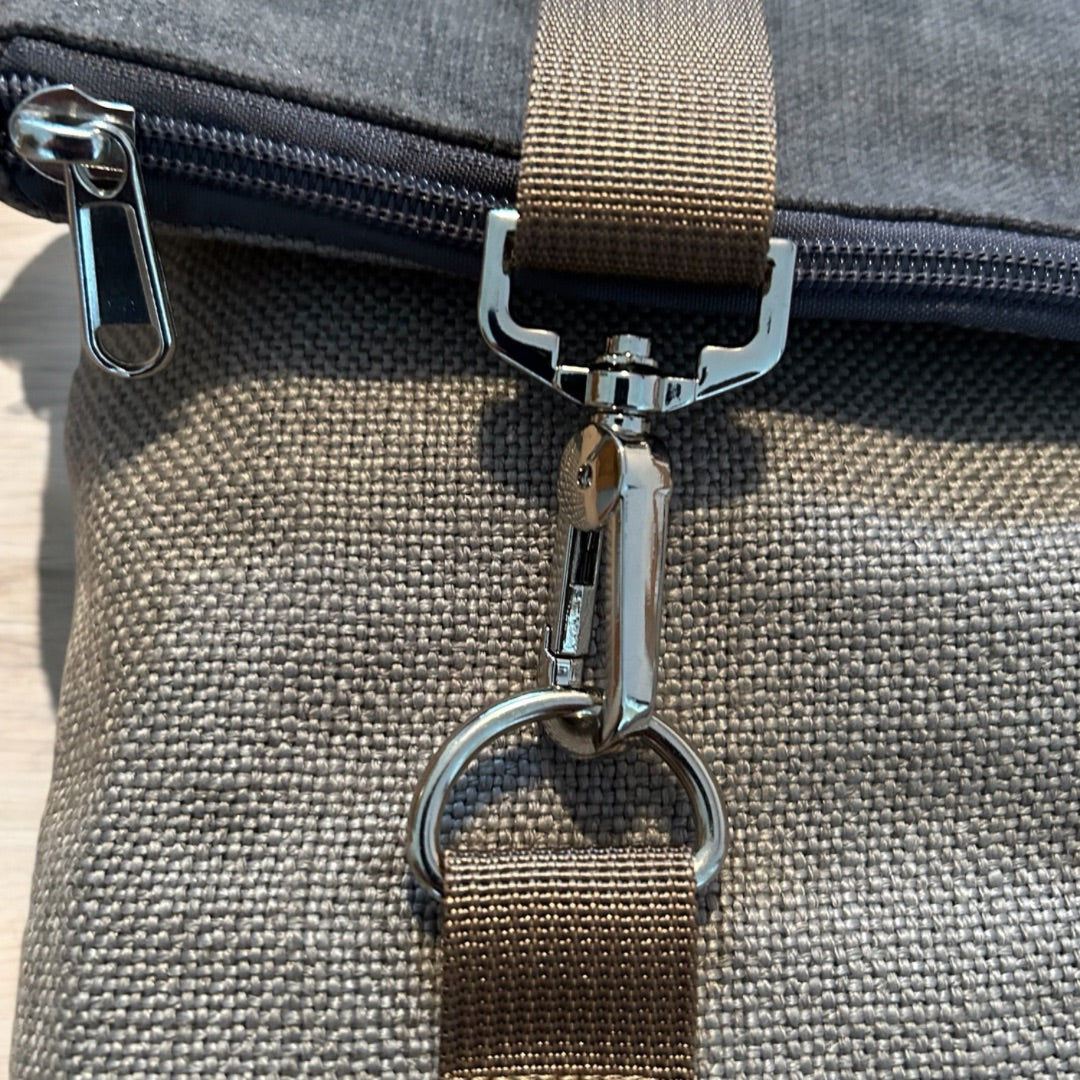 Close-up view of a secure metal clasp and strap attachment on a roll-top backpack. The durable brown strap is fastened to a silver clasp, which connects to a D-ring, ensuring stability and flexibility in the bag's design. The textured fabric of the bag is visible, highlighting its rugged, high-quality material. A zipper with a metal pull tab is seen near the clasp, providing additional functionality and access to the bag's compartments. The sturdy hardware and fabric combination suggest a well-crafted, prac