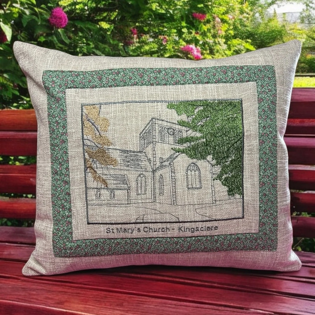 A decorative pillow featuring an embroidered image of St. Mary’s Church in Kingsclere. The design is bordered by a green fabric with small floral patterns, adding a quaint, rustic charm. The pillow has a linen-like texture, and the detailed embroidery includes elements of the church architecture with surrounding trees in green and brown shades. The pillow is displayed on a red wooden bench with a lush garden background filled with greenery and blooming pink flowers, enhancing its cozy and handmade appeal.