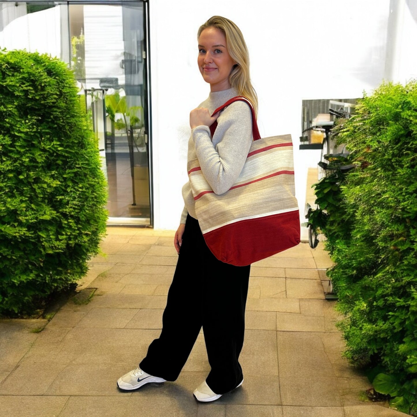 A woman with blonde hair stands outdoors in a paved area surrounded by green shrubbery. She is wearing a light gray sweater, black pants, and white sneakers, smiling slightly at the camera. She carries a large tote bag over her shoulder featuring a beige and red striped pattern with a solid red panel at the bottom. The setting is bright and casual, highlighting the tote bag's earthy and stylish design.