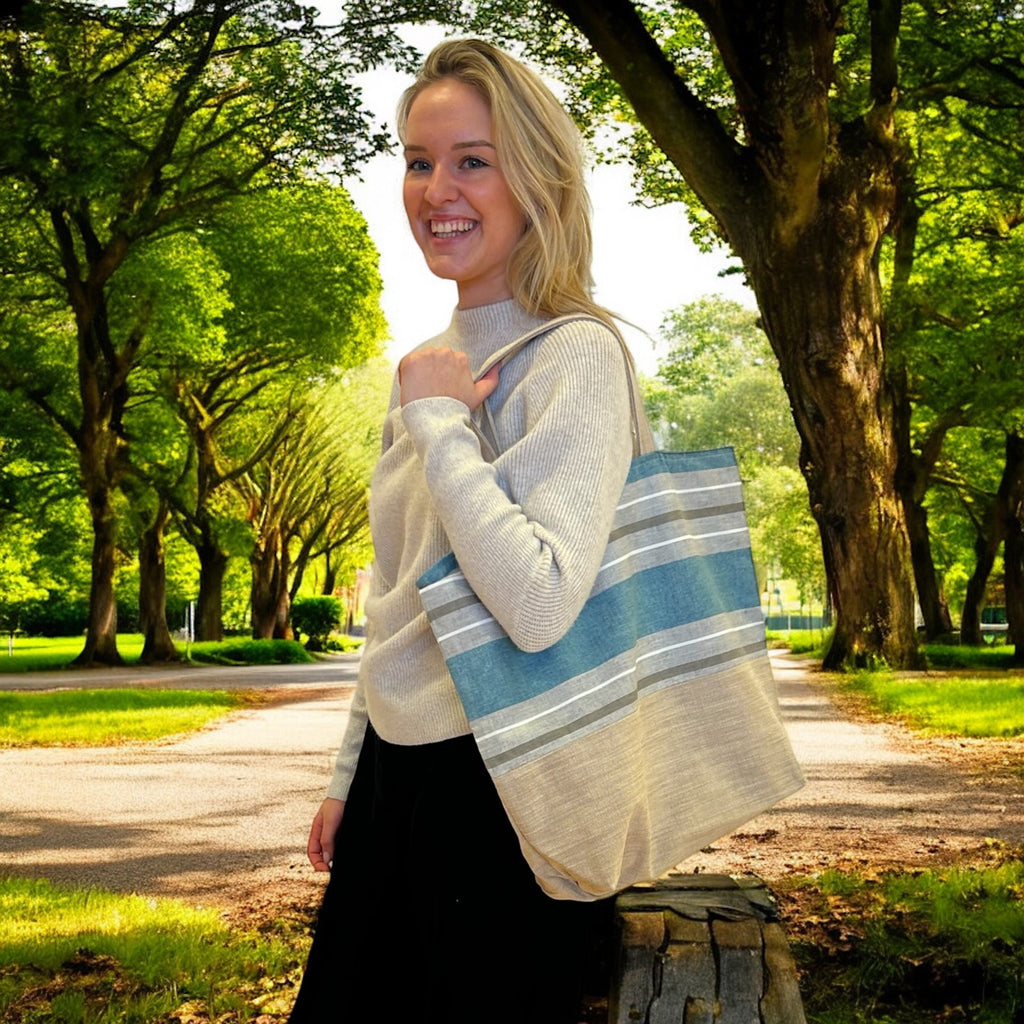 A woman standing outdoors in a sunlit park, smiling while holding a large tote bag on her shoulder. The tote bag features a simple, stylish design with horizontal blue and beige stripes. She is wearing a light-colored sweater and black pants, with trees and a path visible in the background, creating a calm, natural setting.