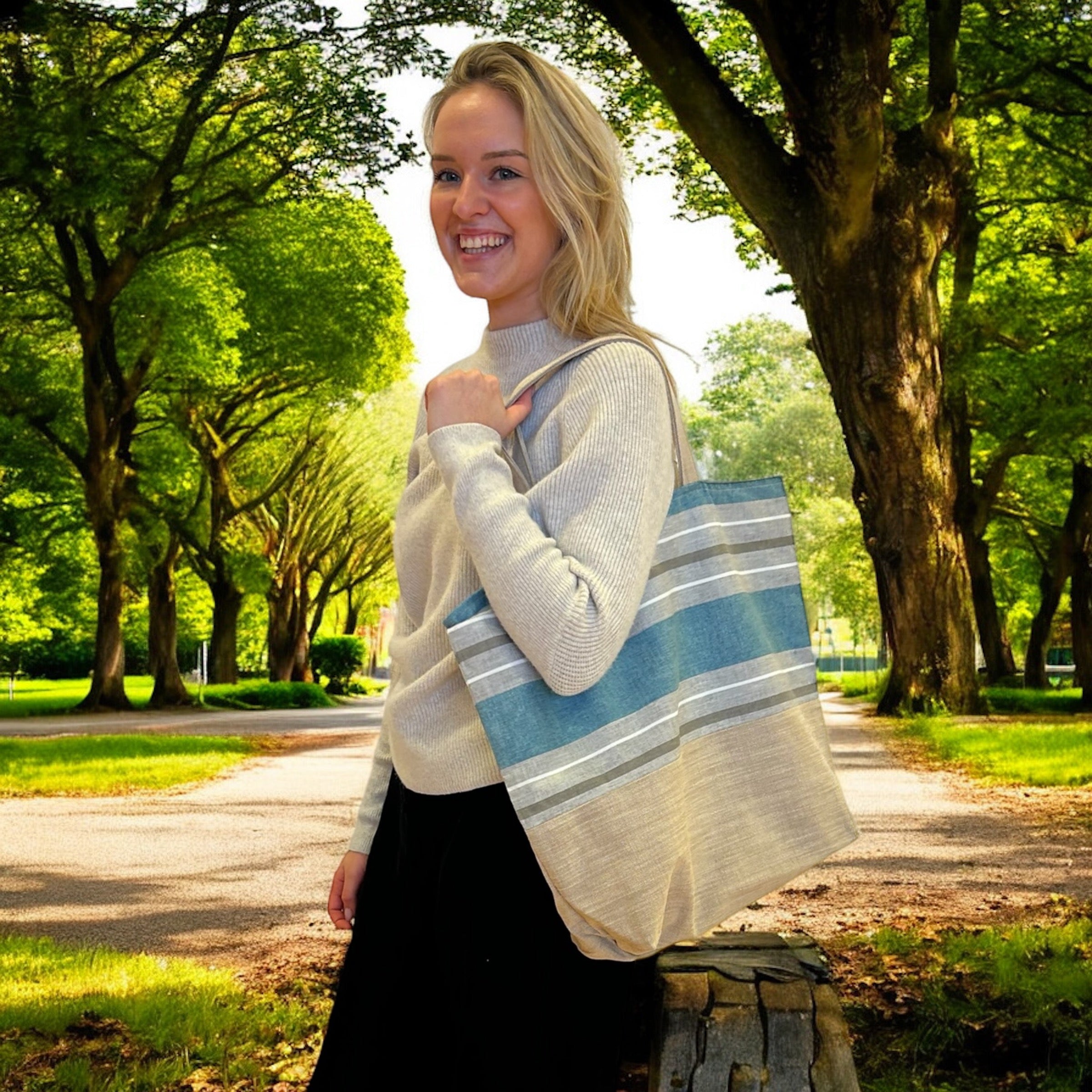A woman standing outdoors in a sunlit park, smiling while holding a large tote bag on her shoulder. The tote bag features a simple, stylish design with horizontal blue and beige stripes. She is wearing a light-colored sweater and black pants, with trees and a path visible in the background, creating a calm, natural setting.