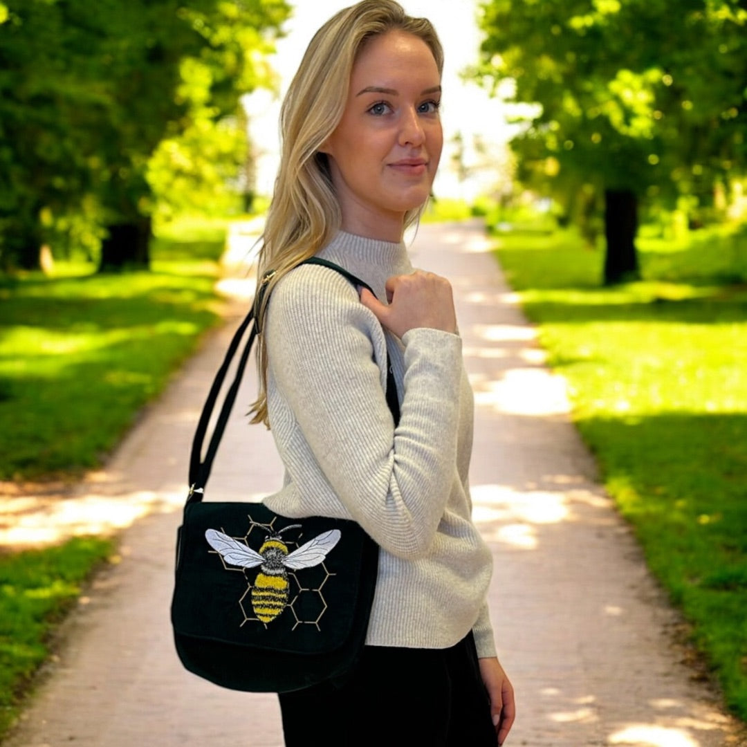 A woman with blonde hair stands on a sunlit pathway lined with lush green trees, holding a black shoulder bag with an embroidered bee design on the flap. The bee is detailed in yellow, white, and black, set against a background of golden honeycomb outlines. She is wearing a light gray sweater and smiles subtly at the camera, creating a relaxed and natural look. The vibrant greenery and bright light add to the fresh, outdoor atmosphere.