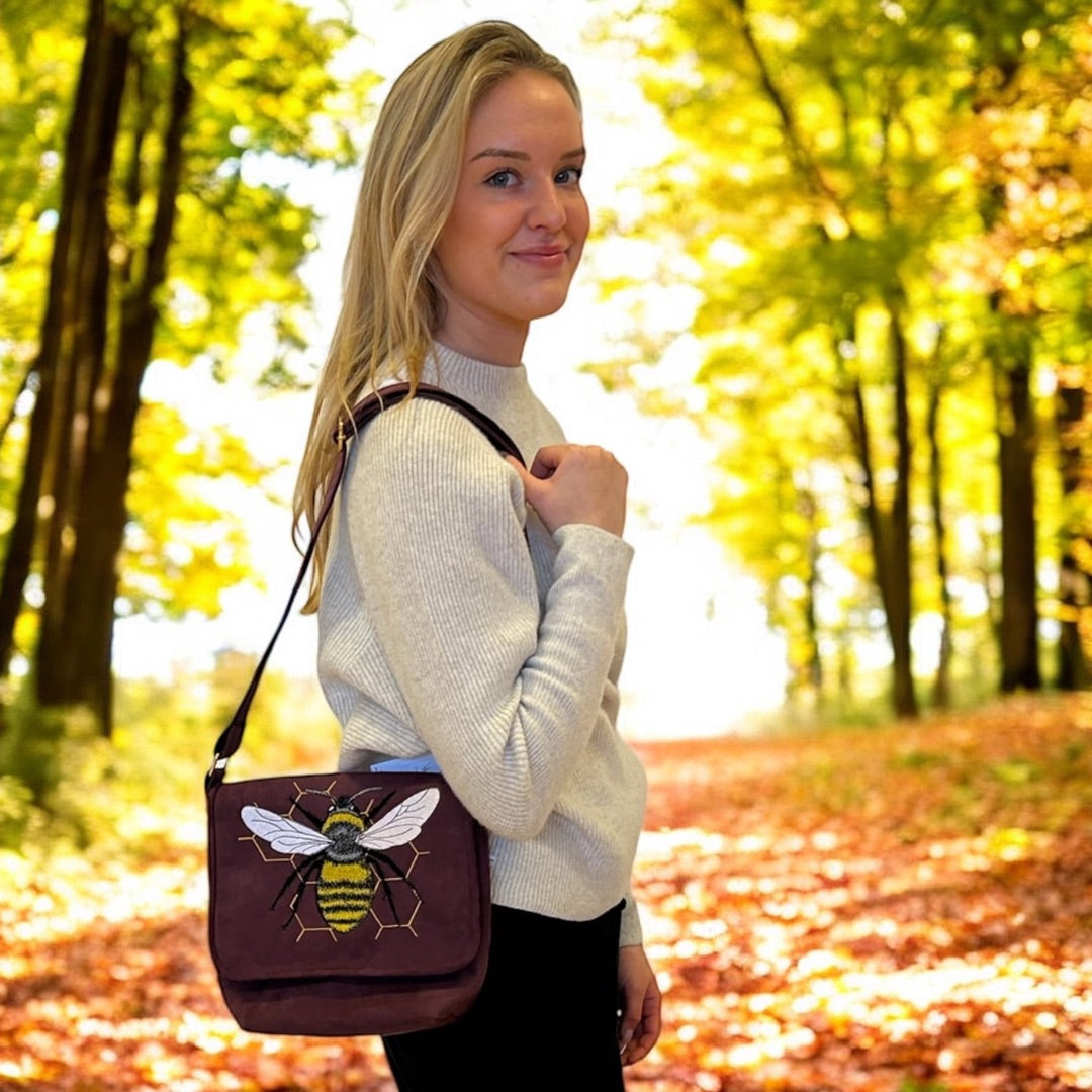 A woman with blonde hair stands on a leaf-strewn path in a sunlit forest during autumn. She is wearing a light gray sweater and holding a maroon shoulder bag with an embroidered design of a bee on the flap. The bag's detailed bee design features yellow, black, and white stitching, with a honeycomb pattern in the background. The woman's subtle smile and the warm autumn colors create a cozy, nature-inspired atmosphere.