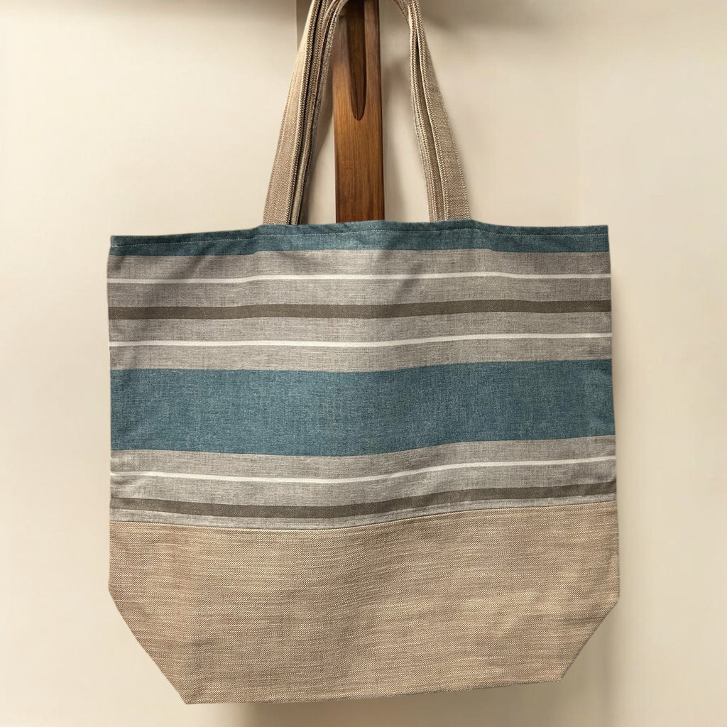 A large tote bag hanging on a wooden peg against a plain background. The bag features a simple design with horizontal stripes in shades of blue, beige, and gray, with a textured fabric. It has sturdy fabric handles and a spacious, boxy shape, making it ideal for everyday use.