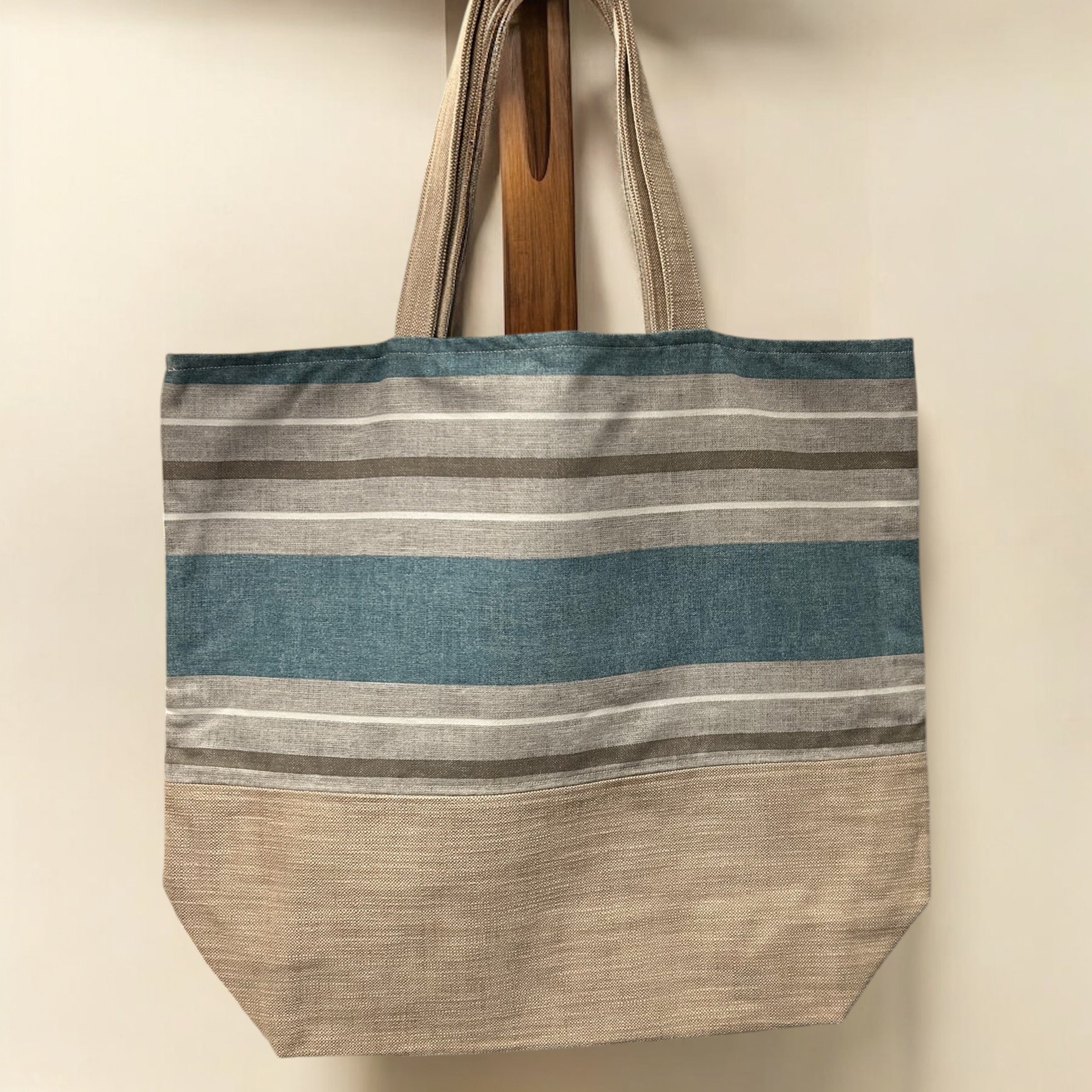 A large tote bag hanging on a wooden peg against a plain background. The bag features a simple design with horizontal stripes in shades of blue, beige, and gray, with a textured fabric. It has sturdy fabric handles and a spacious, boxy shape, making it ideal for everyday use.