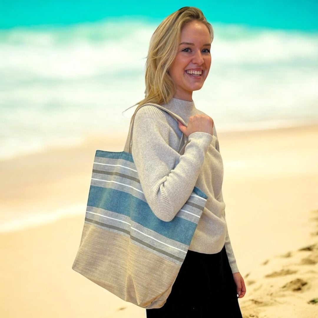 A woman smiling on a sandy beach, holding a large tote bag with blue and beige horizontal stripes on her shoulder. She is wearing a light-colored sweater and black pants, with the ocean and waves in the background, creating a relaxed and sunny coastal scene.