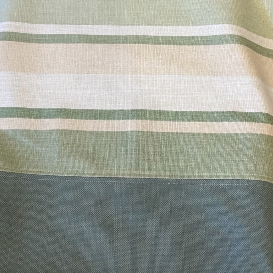 A close-up view of the fabric on a tote bag, showcasing a soft green, cream, and beige striped pattern. The lower section features a solid green panel with a slightly textured finish. The fabric has a natural, woven appearance, highlighting the bag's quality and understated, earthy color palette.