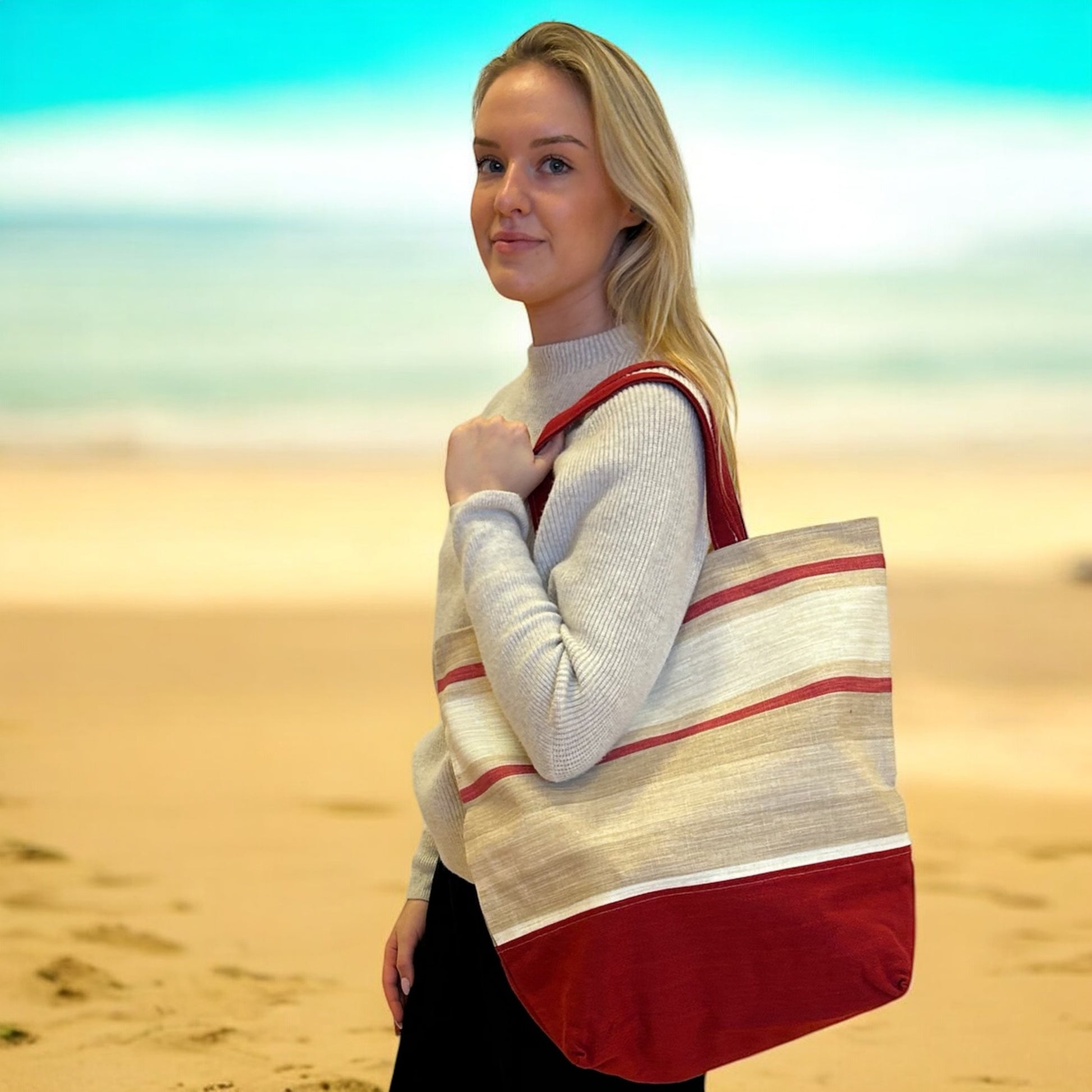 A woman with blonde hair stands on a beach with sand and a blurred ocean backdrop, holding a large tote bag over her shoulder. She is wearing a light gray sweater. The tote bag features a red and beige striped pattern, with a solid red panel at the bottom. The bag has a natural, earthy look that complements the beach setting. The woman looks at the camera with a slight smile, creating a relaxed and serene atmosphere.
