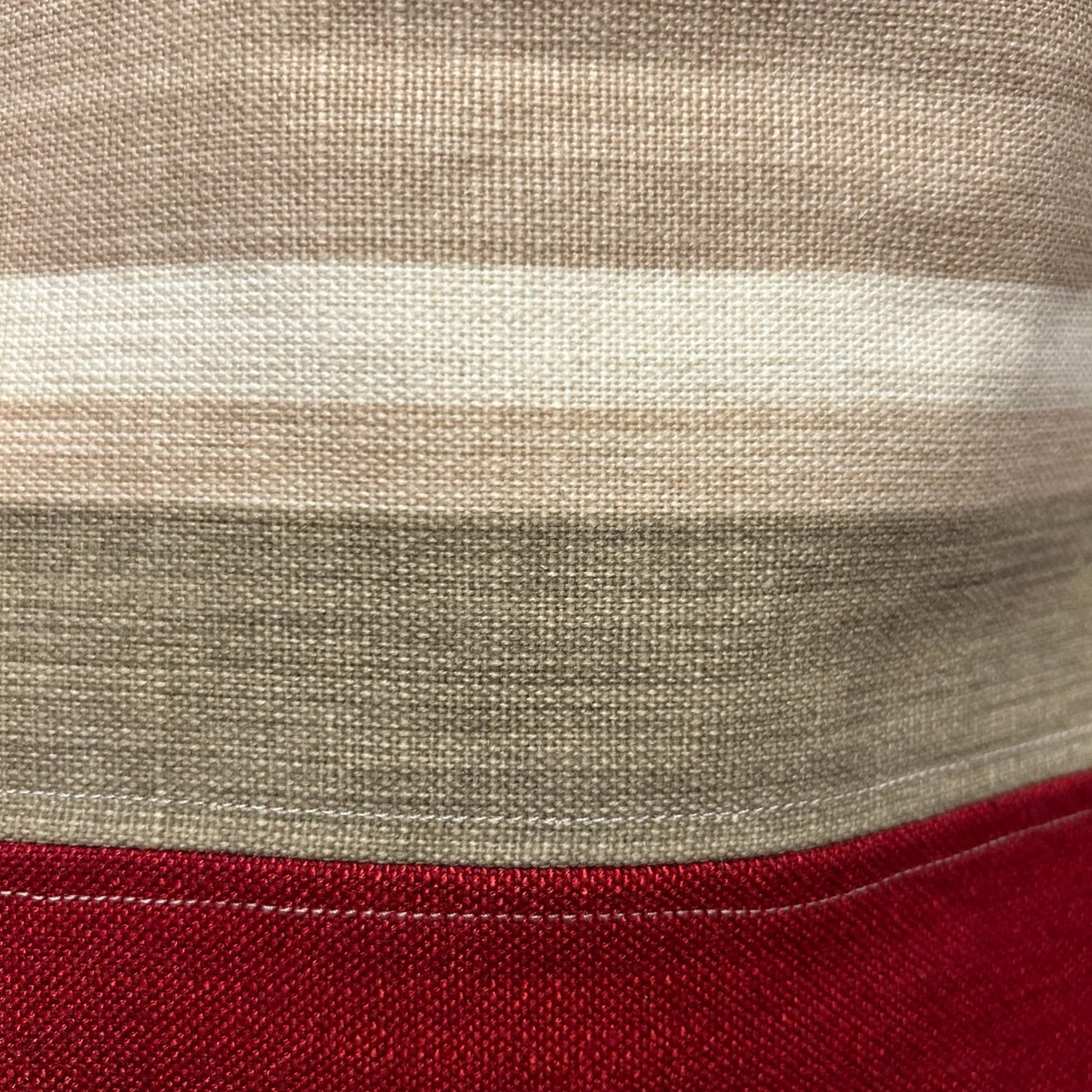 A close-up view of the fabric on a tote bag, showcasing a striped design in shades of beige, cream, green, and red. The texture of the woven fabric is clearly visible, adding a rustic and natural feel to the material. The bottom section features a solid red panel with visible stitching, enhancing the handcrafted look of the bag.
