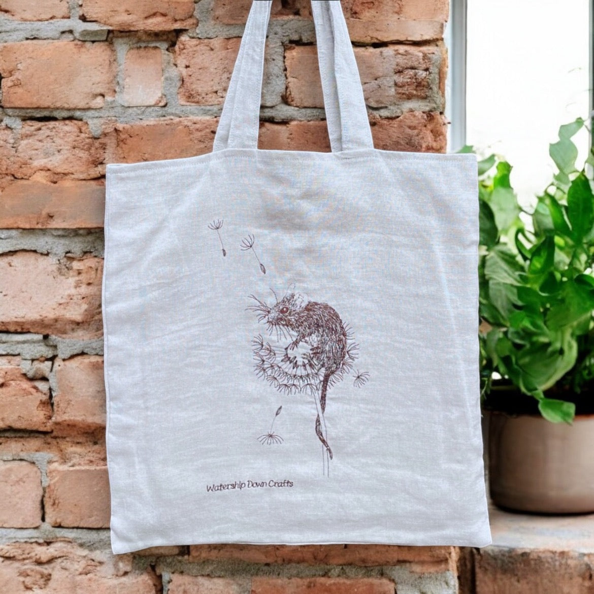 Eco-friendly handmade linen tote bag with brown embroidered design of a mouse on a dandelion, by Watership Down Crafts. This unique, nature-inspired tote features intricate details of a mouse perched on a dandelion with seeds floating, perfect for animal and nature lovers. Lightweight, reusable, and ideal as a sustainable gift or everyday bag, displayed against a rustic brick wall with green plants in the background.