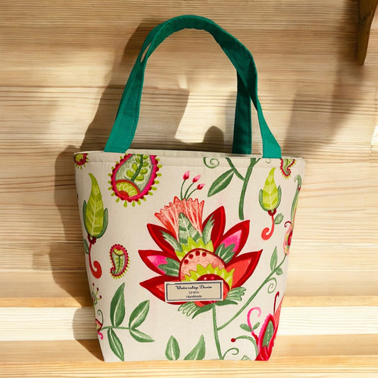 A handmade mini tote bag featuring a bold and vibrant floral design with bright red, green, and pink patterns on a neutral beige background. The bag is accented with teal handles that add a fresh pop of color and a "Watership Down Crafts Handmade" label on the front. It is displayed on a light wooden shelf, illuminated by natural sunlight, showcasing its cheerful and artistic style.