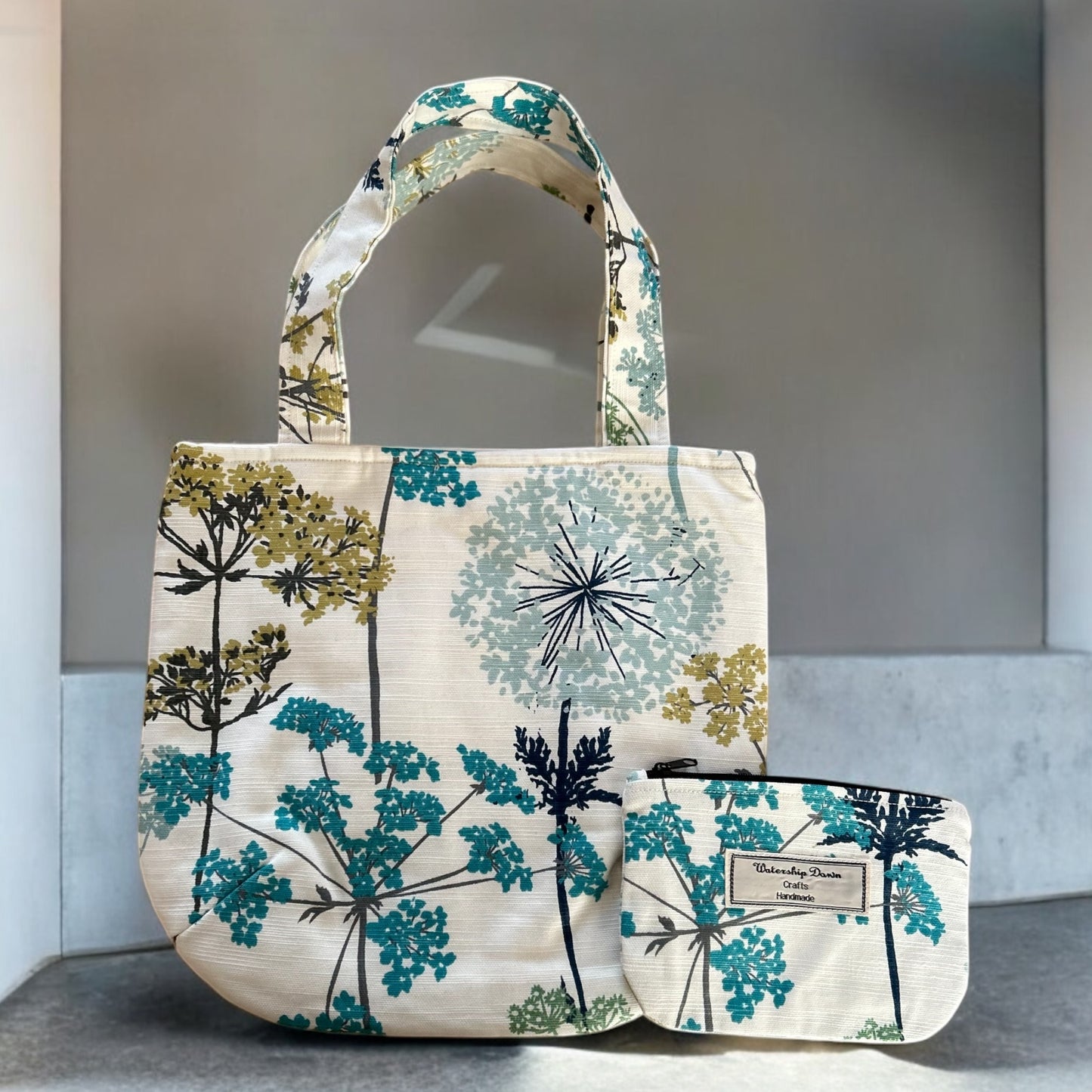 A stylish handmade handbag tote bag and matching pouch featuring a botanical print with large dandelion-like flowers in shades of blue, teal, and mustard yellow. The tote bag has sturdy handles and a practical, spacious design, while the smaller pouch complements it with the same floral pattern. Both items prominently display the "Watership Down Crafts Handmade" label, emphasizing their handcrafted quality. The set is photographed against a minimalist background, highlighting the elegant and modern design. 