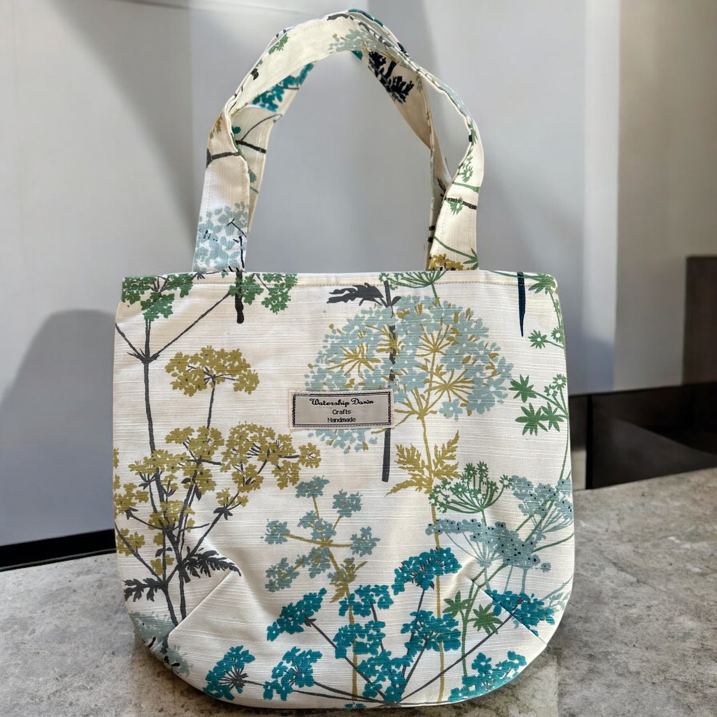 A handmade mini tote bag featuring a beautiful botanical print with large, delicate flowers in shades of teal, blue, and mustard yellow. The design showcases abstract floral elements against a soft cream background, giving the bag a fresh, nature-inspired aesthetic. The "Watership Down Crafts Handmade" label is prominently displayed on the front, emphasizing the bag’s handcrafted quality. It is photographed in a minimalist setting, highlighting its modern and elegant look.