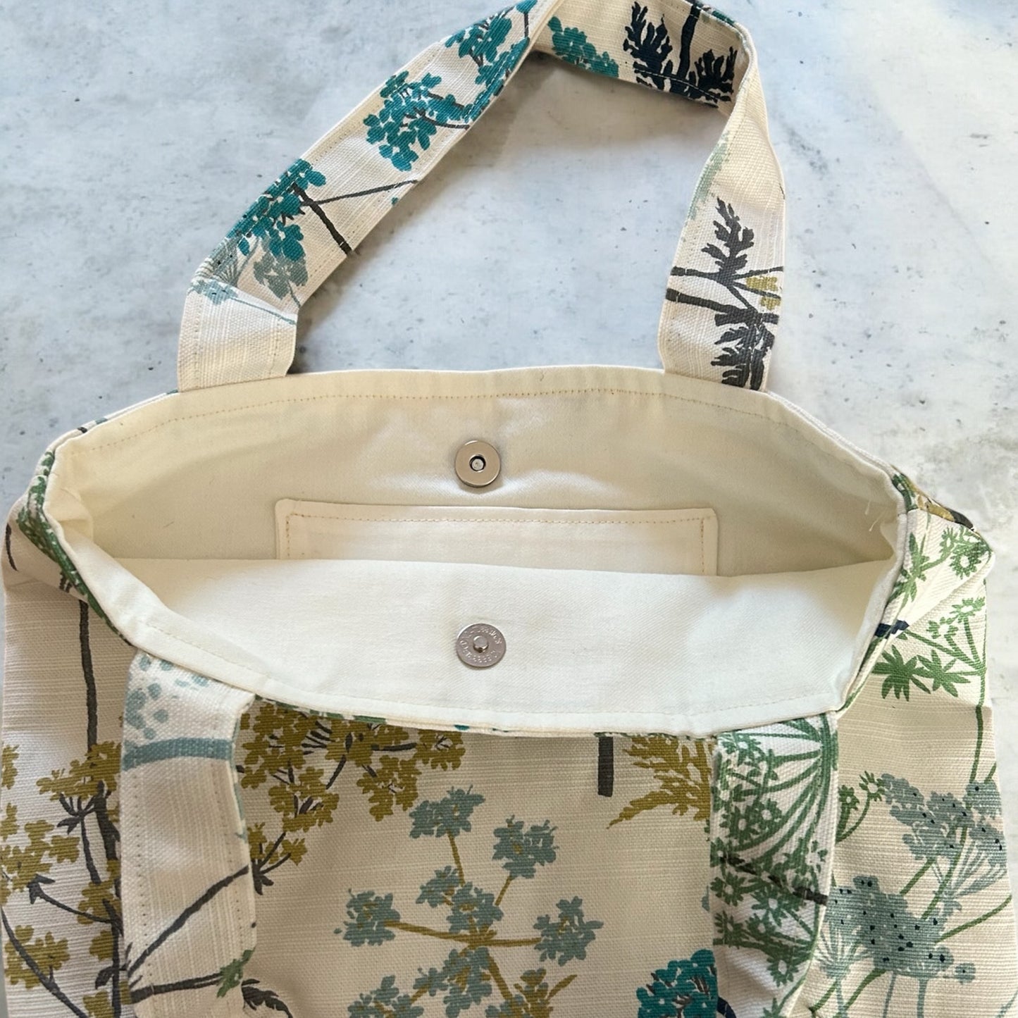 A close-up of the interior of a handmade tote bag, showing a clean and simple lining with a magnetic snap closure for secure fastening. The inner fabric is a solid beige color, complementing the bag's botanical print on the outside. The "Watership Down Crafts Handmade" label is also visible on the inside, emphasizing the bag's handcrafted quality. The design features a practical, minimalist style with functional details.