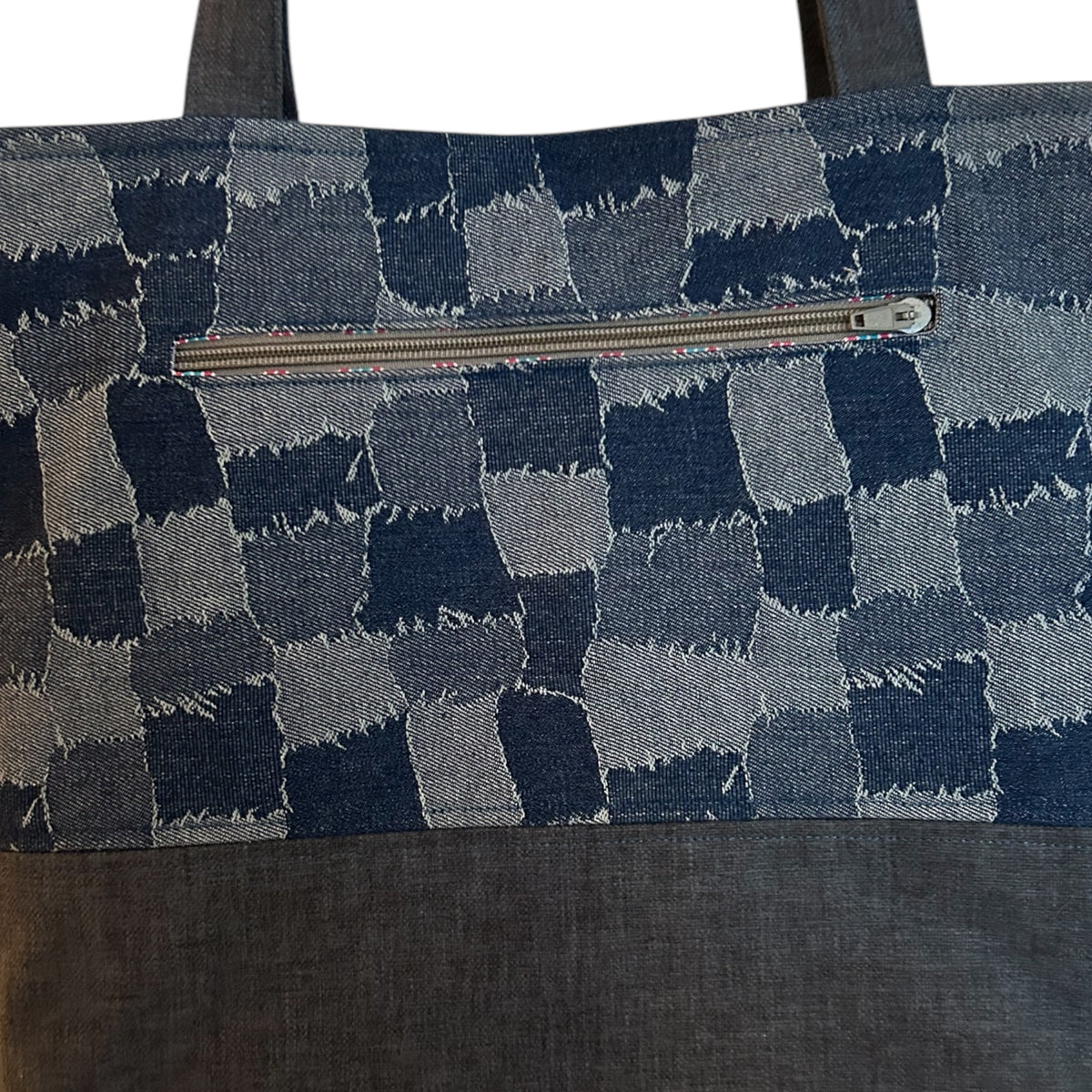 Close-up of a denim patchwork tote bag featuring a horizontal zippered pocket with intricate stitching and a striped fabric lining. The checkerboard design highlights its handcrafted appeal, combining style and functionality.