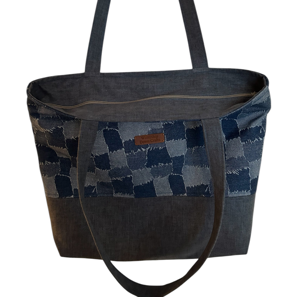 Close-up view of a denim patchwork tote bag featuring a checkerboard design with frayed edges, long straps, and a durable, structured base. The bag includes a leather 'Watership Down Crafts' logo tag, showcasing its handmade quality and stylish functionalit