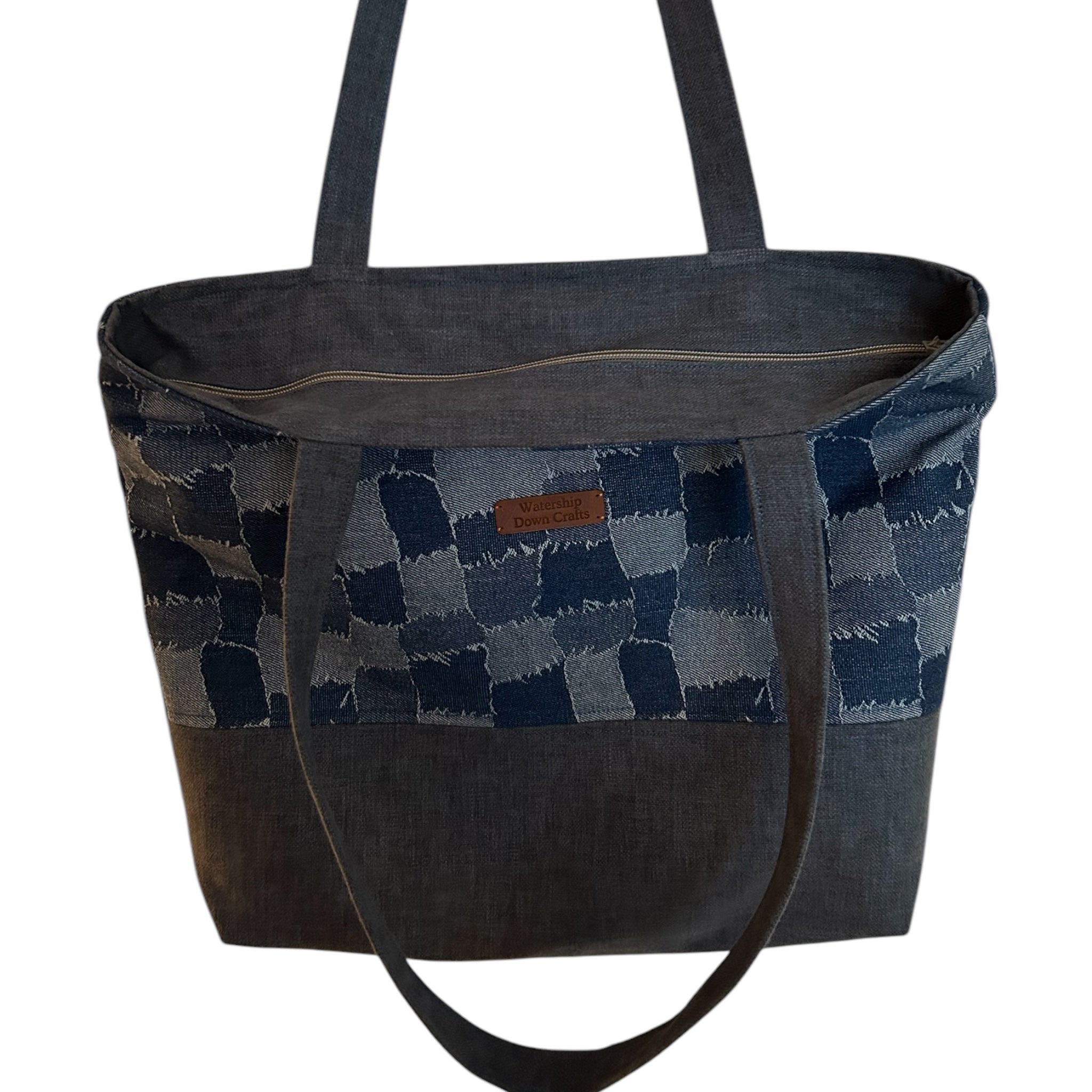 Close-up view of a denim patchwork tote bag featuring a checkerboard design with frayed edges, long straps, and a durable, structured base. The bag includes a leather 'Watership Down Crafts' logo tag, showcasing its handmade quality and stylish functionalit