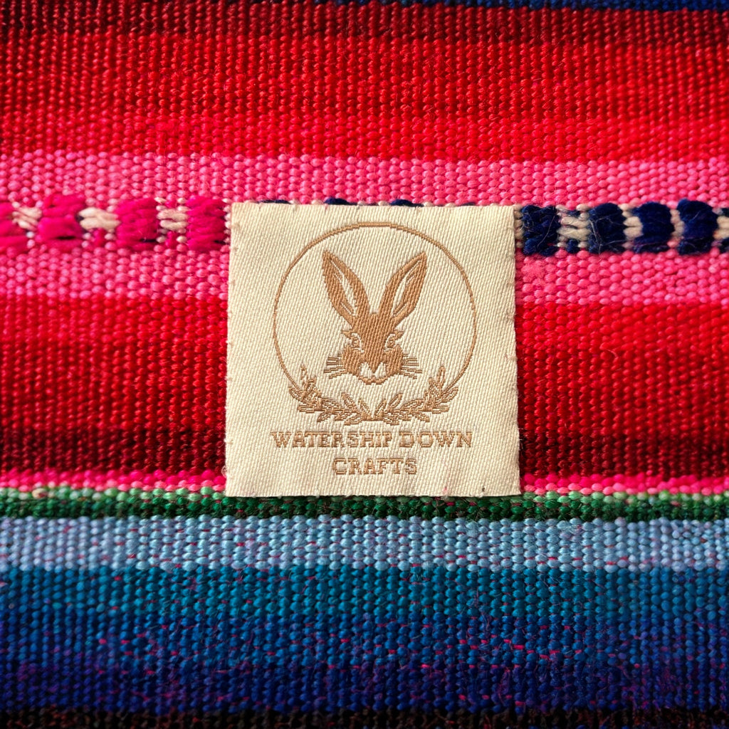 Close-up of the Watership Down Crafts logo tag on a vibrant pink, blue, and green woven fabric, showcasing the fine craftsmanship and unique branding of this handmade product