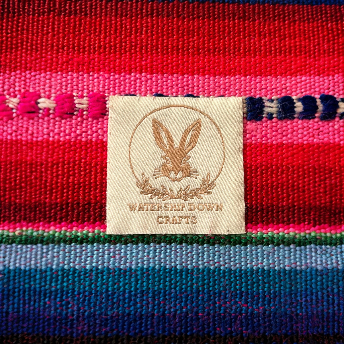 Close-up of the Watership Down Crafts logo tag on a vibrant pink, blue, and green woven fabric, showcasing the fine craftsmanship and unique branding of this handmade product
