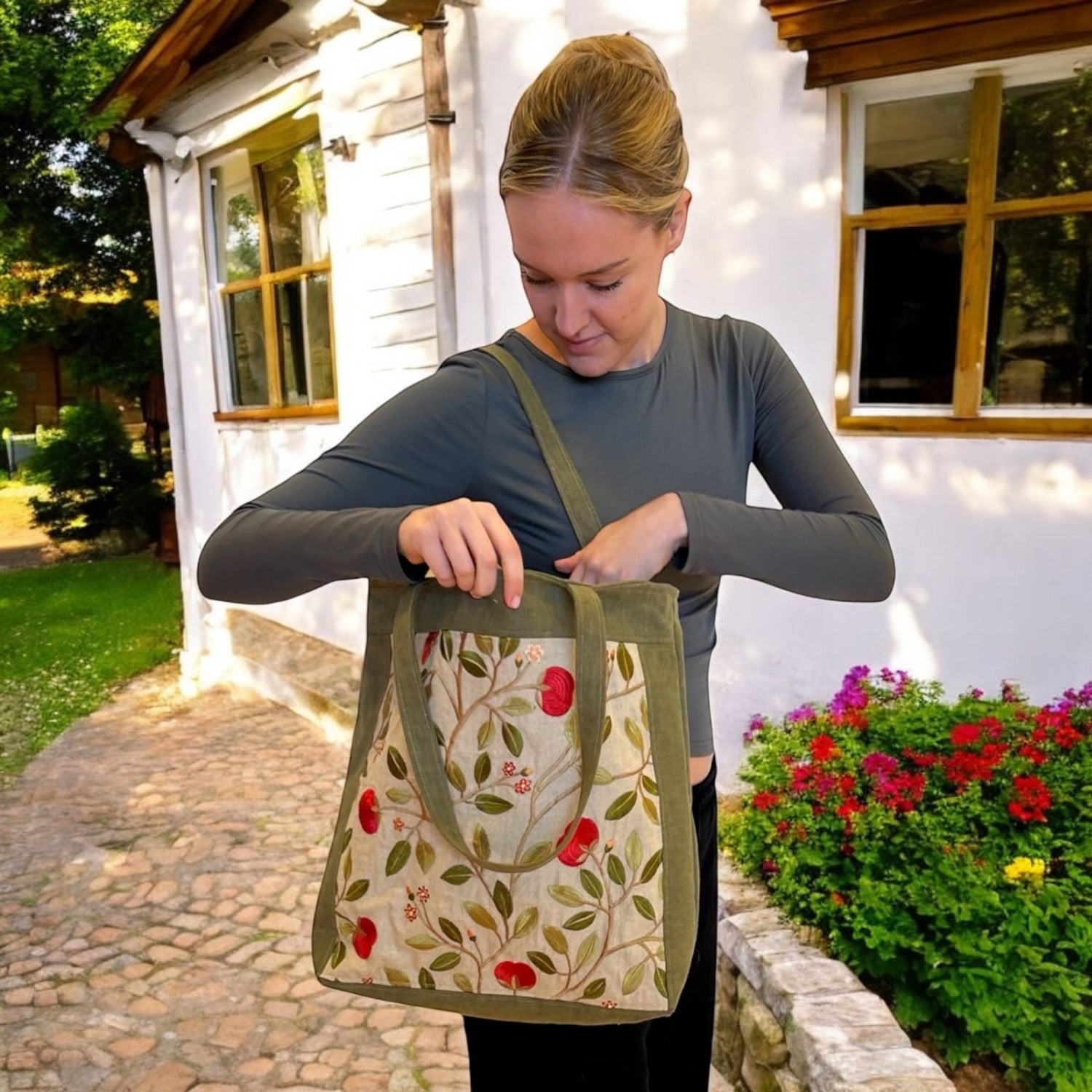 A woman standing outdoors holding a beige and green embroidered tote bag with a floral pattern and zip closure.