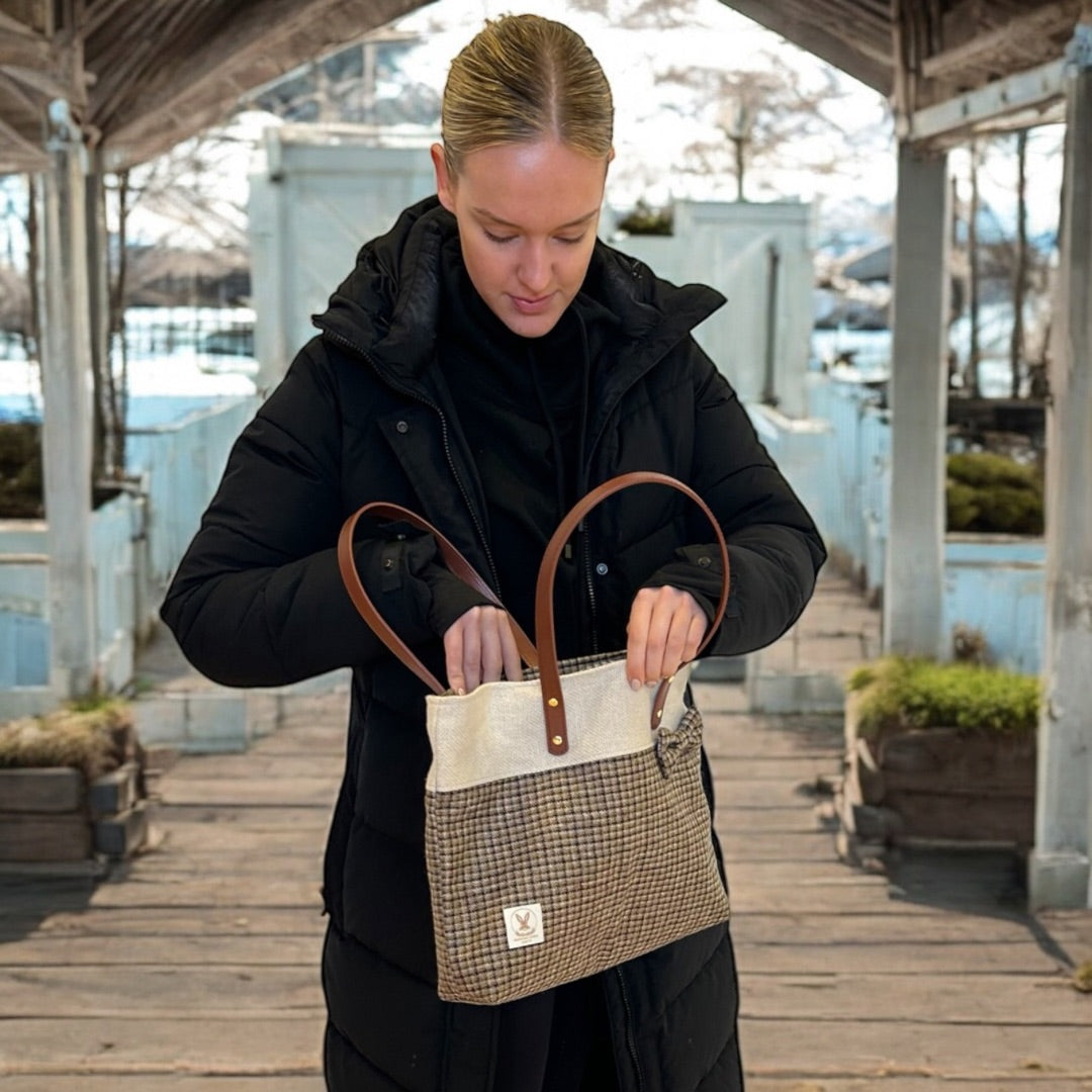 Woman in a black coat standing outdoors, looking into a tweed tote bag with leather handles.