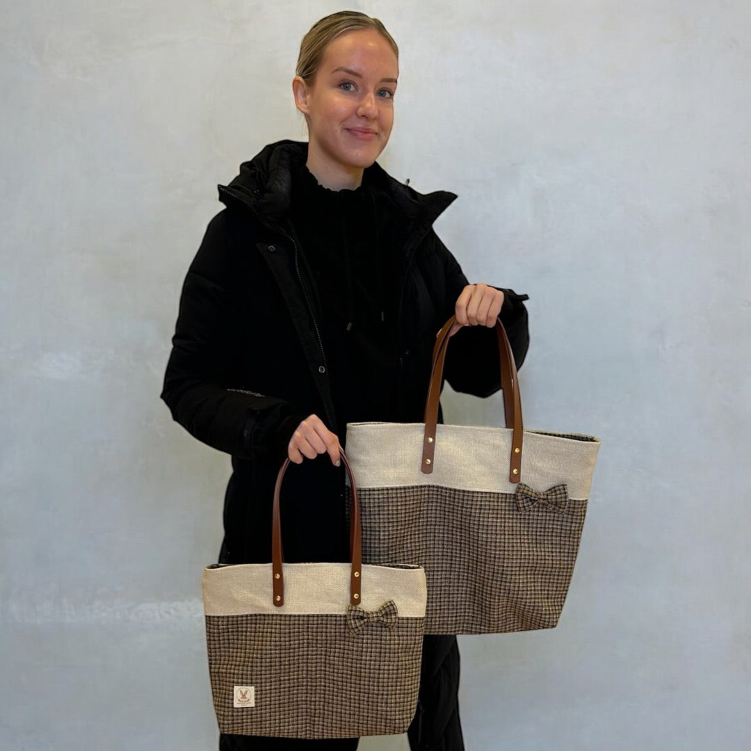 Smiling woman in a black coat holding two tweed tote bags with leather handles and bow details.